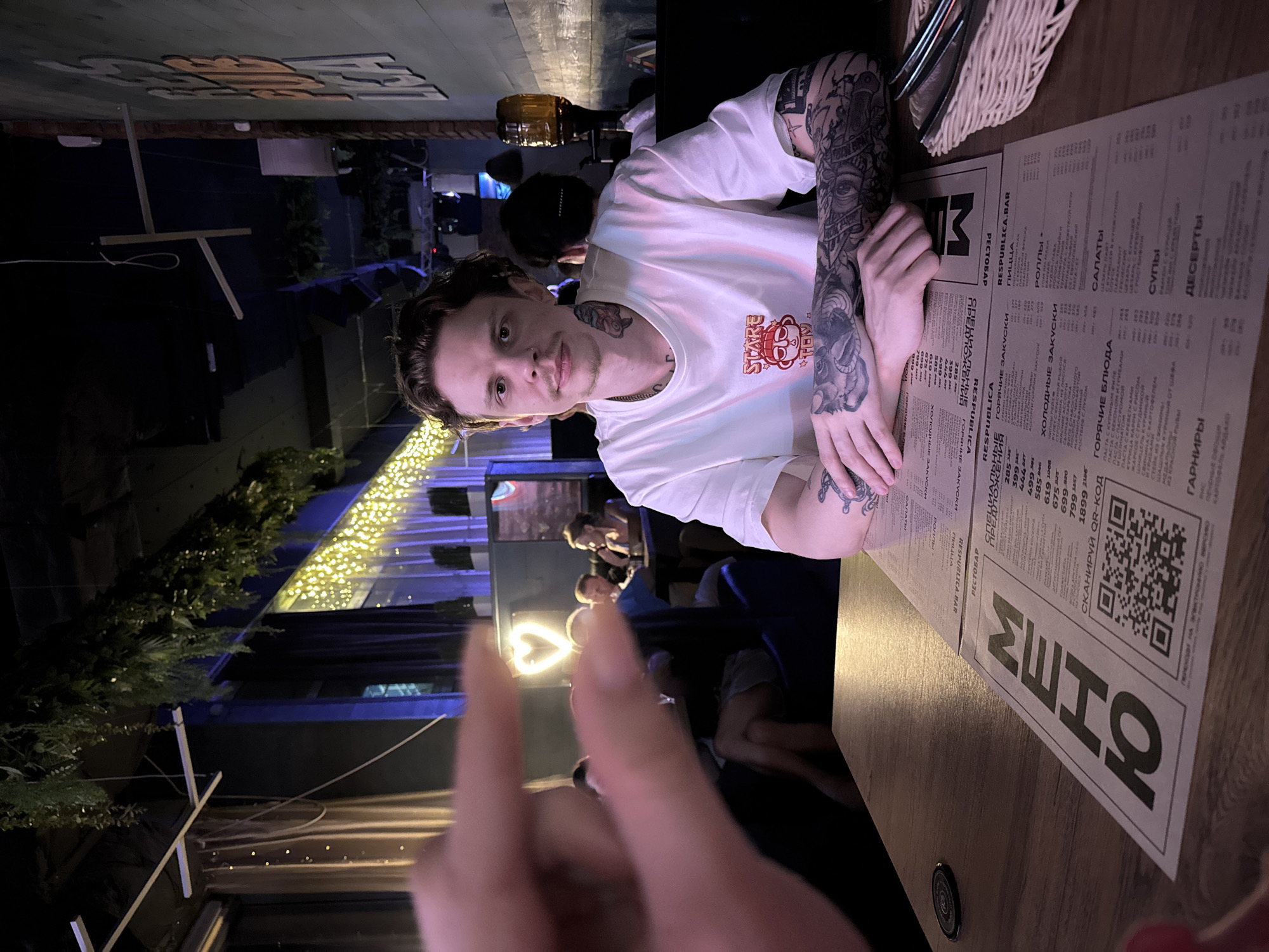 The image shows a young man with tattoos on his neck and arms, wearing a white t-shirt, sitting at a table in a restaurant. He is looking directly at the camera. In the foreground, a blurred hand is positioned as if indicating something. A menu is laid out on the table in front of him, with a QR code visible. The restaurant appears to be dimly lit with string lights and hanging plants, suggesting an evening or indoor setting. In the background, other patrons are visible, some seated at tables. A large beer dispenser is present on a shelf in the background. The overall atmosphere seems casual and social.
