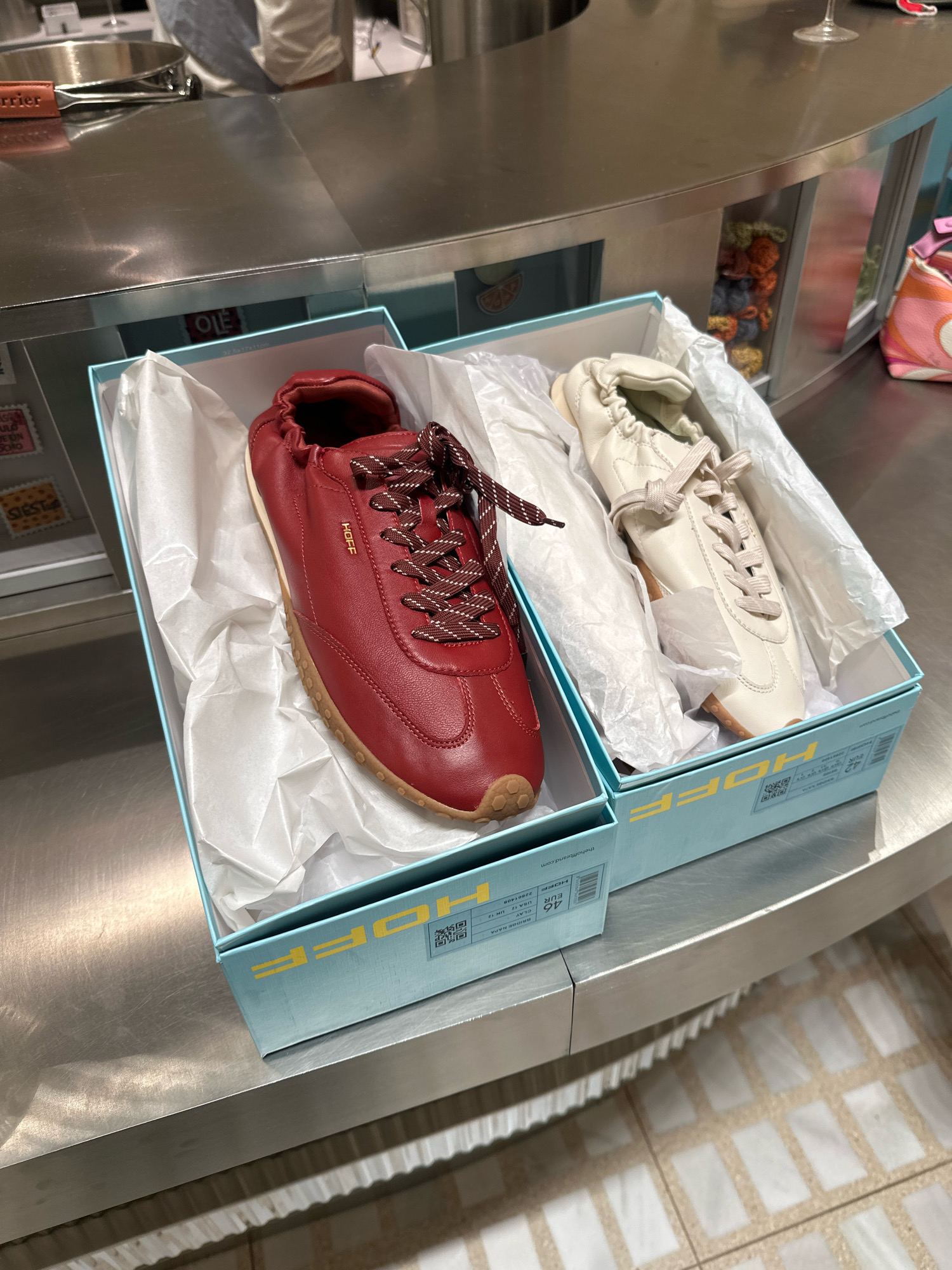 Two shoeboxes, one light blue and one slightly larger light blue, are placed side-by-side on a stainless steel counter. The boxes are open, revealing pairs of sneakers. The left box contains a pair of burgundy sneakers with white and burgundy checkered laces, and a gum-colored sole. The right box contains a pair of off-white sneakers with off-white laces and a similar gum-colored sole. Both pairs of sneakers have elastic ruched tops. The boxes are branded with the word "HOFF" in yellow lettering on the sides, and some product information including what appears to be "thehoffbrand.com". Behind the shoeboxes, the stainless steel counter continues, with some reflective surfaces. In the background, a blurred figure of a person wearing a white shirt and a grey apron can be seen behind a curved counter. To the right of the shoeboxes, a small pile of colorful knitted objects is visible through a glass partition. Further to the right, a pink and orange patterned handbag rests on the counter. The floor appears to be tiled with a grid pattern. The scene suggests a retail environment, possibly a shoe store or a boutique in Madrid, Spain, given the mention of "Madrid" and "Spain" in the prompt.