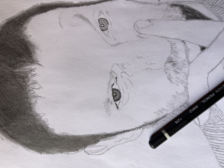 The image presents a close-up view of a monochrome pencil sketch of a man's face on white paper, taken in the city of Jos, Nigeria. The drawing depicts the man from the top of his head, showing dark, textured hair, down to his upper chest. He features a mustache and a beard, rendered with distinct pencil strokes. Both of the man's eyes are open, with detailed pupils and irises, and gaze generally forward. His right index finger is positioned vertically, touching his upper lip and the base of his nose, suggesting a gesture of quiet or contemplation. Partially visible on the lower left of the drawing are vertical lines indicating part of a striped garment or collar. A black 'SKETCH PENCIL 8802 12B' pencil rests diagonally across the lower left quadrant of the drawing, its tip pointing towards the man's face. The drawing surface is unlined white paper, with minor smudges visible around the graphite rendering.