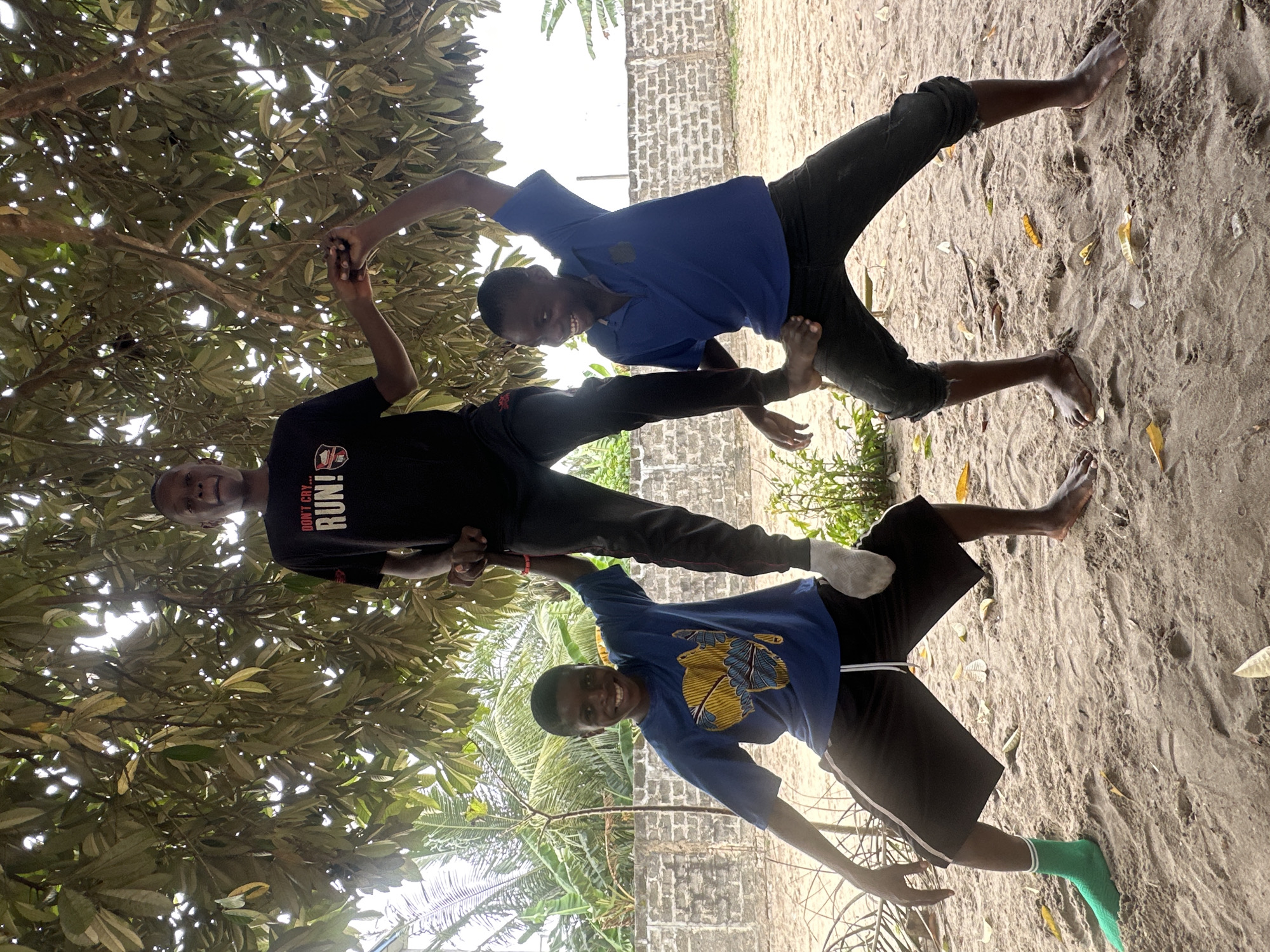 Three young males are posing for a photo in an outdoor setting, possibly in the city of Badagry, Nigeria. The person in the middle is standing on the shoulders of the person on the left, and they are both supporting the person on the right, who is holding onto the person in the middle's arms and head. The person in the middle is wearing a black t-shirt with white text that reads "DON'T CRY! RUN!". The person on the left is wearing a blue t-shirt with a yellow and blue graphic of Africa on it, and black shorts. They are also wearing a green sock on their left foot. The person on the right is wearing a blue polo shirt and black pants. They are smiling and looking at the camera. The background shows trees, foliage, and a brick wall. The ground is sandy.