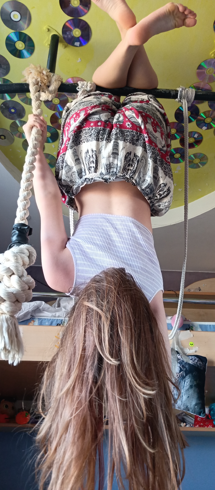 A child is hanging upside down from a black metal bar, with their feet crossed at the ankles and resting on the bar. They are wearing a red and white patterned, elasticated, midriff-baring top and a white, vertically striped top that is riding up their back. The child is holding onto a thick rope with their left hand. Their brown hair hangs downwards.
The background consists of a yellow ceiling covered in affixed CDs. A bed with a light-colored mattress is partially visible behind the child, with a shelf containing various objects including a dark-colored pillow, visible behind that. The location is Unknown, Unknown.
