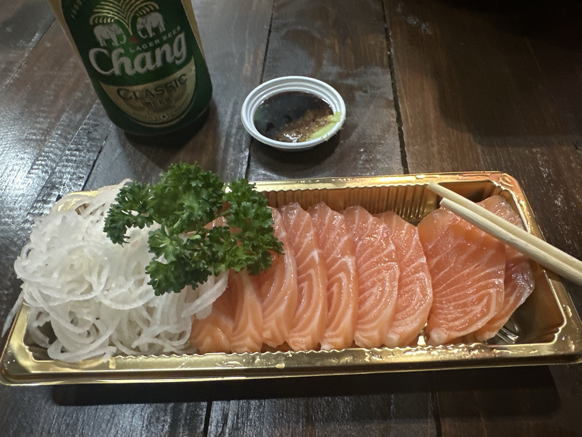 The image captures a scene of dining in Pattaya City, Thailand. Centered is a golden rectangular tray filled with salmon sashimi, accompanied by shredded daikon radish, parsley, and a pair of chopsticks resting on the edge. To the left of the tray, there's a bottle of Chang Classic beer and a small bowl of soy sauce with wasabi. The setting is a wooden table.
