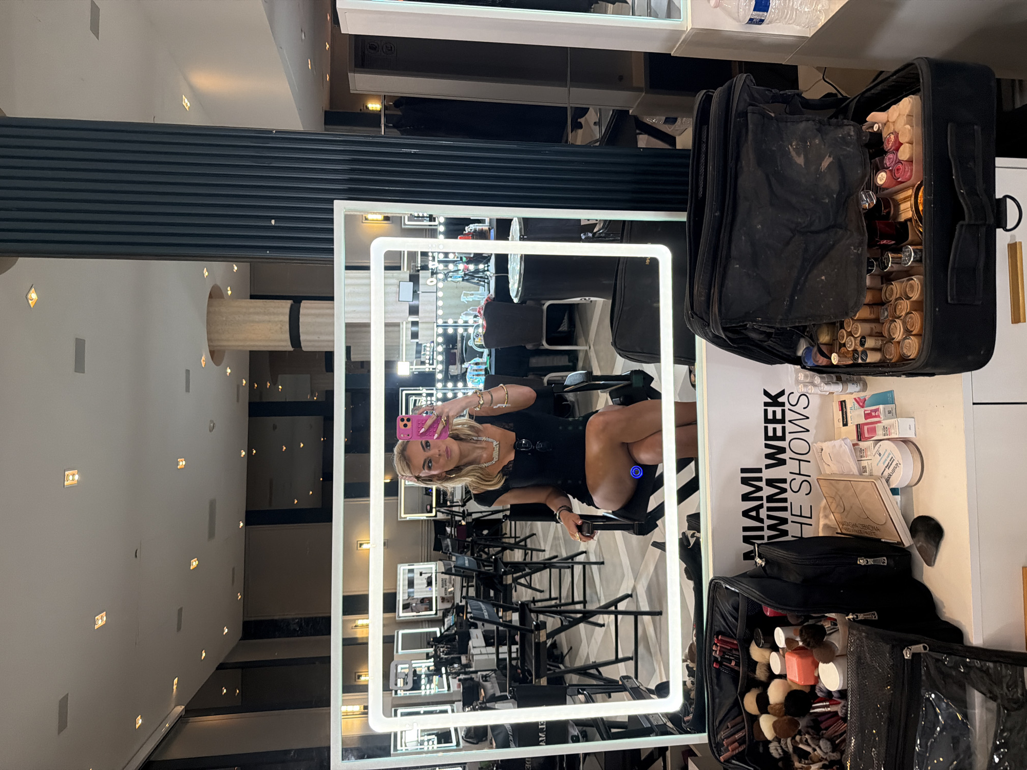 The image shows a woman taking a selfie in a mirror at what appears to be a makeup station for Miami Swim Week, as indicated by the text on a nearby sign. The location is likely in Miami Beach, Florida, within a building featuring columns and architectural details. The woman is seated in a chair, holding a pink phone up to take the photo. Several similar makeup stations with mirrors and chairs are visible in the background. Makeup supplies and equipment are seen on the table next to the mirror.