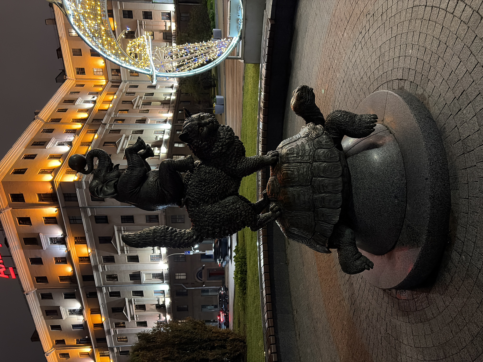 A bronze sculpture depicts a composition of animals: a turtle at the base, supporting a wolf-like creature, which in turn supports two elephants balancing a ball. The sculpture stands on a round pedestal, set on a pavement of gray cobblestone arranged in an arc pattern. Behind the sculpture, there is a narrow strip of green lawn bordered by a low retaining wall made of dark bricks. A multi-story building with lit windows forms the backdrop. A decorative light structure, possibly part of city decorations, is visible above the building. A red vertical sign with visible letters is attached to the building. The scene is lit, suggesting it is nighttime. The location is likely in a city, although the specific details are not discernible from the image to identify the exact location.