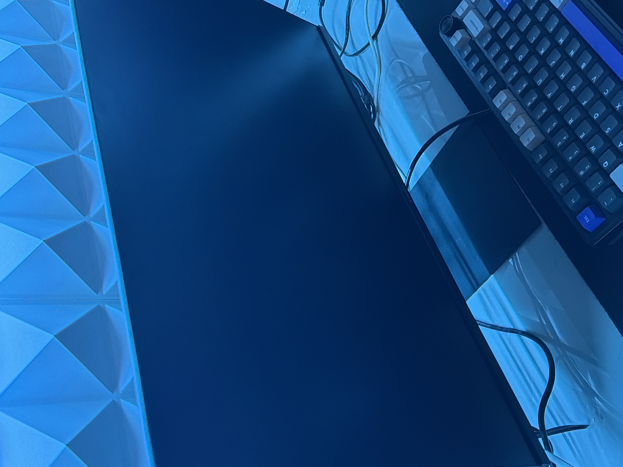 The image shows a close-up of a dark computer monitor placed in front of a geometric, illuminated blue wall panel. Below the monitor, a black keyboard with blue and white keycaps is visible, positioned on a reflective surface. Several black cables are draped across the scene. The image is a still photograph and does not depict any people, actions, or interactions. There is no information available to suggest this scene is located in Wonoayu, Indonesia.
