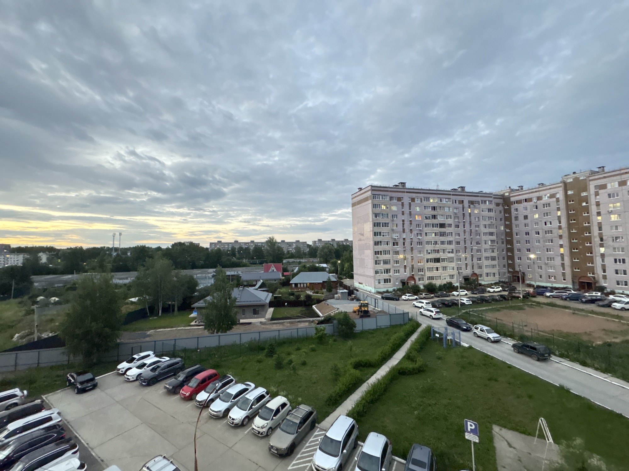 The image depicts a wide-angle, high-angle view of a residential area in Chaikovsky, Russia, likely taken during overcast twilight conditions, evidenced by the gray, cloudy sky with hints of yellow on the horizon.
In the foreground, a parking lot is filled with numerous cars. To the left, a long row of parked cars is visible, mostly white and black. Further into the lot, a smaller group of cars includes a black SUV, two white sedans, a dark gray SUV, a red sedan, and several other sedans and SUVs in white, silver, and gray.
Beyond the parking lot, a road winds through a grassy area. Several cars are parked along this road and in the wider street in front of apartment buildings. One dark green SUV is visible driving on the road.
Dominating the right side of the image are several multi-story apartment buildings, typical of Soviet-era architecture, with numerous windows, some of which are lit. In the distance, more apartment buildings and some smaller houses with red roofs are visible, interspersed with trees and vegetation. A construction site with yellow heavy machinery is also present in the middle ground. A fence runs along the edge of the grassy area, separating it from the residential structures. A prominent blue parking sign is visible in the lower right quadrant of the image.