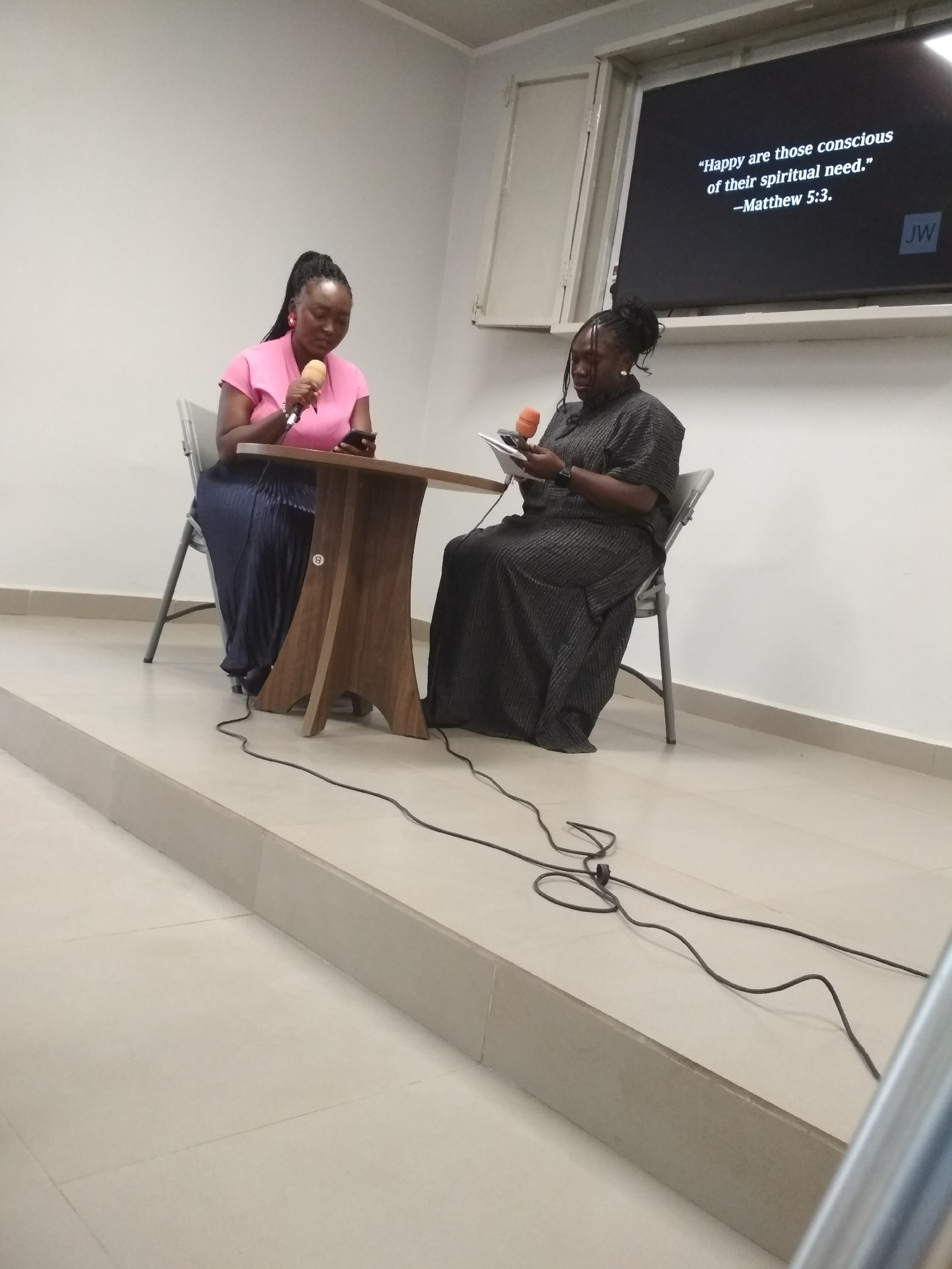 Two women are seated at a wooden table on a low platform, both holding microphones and looking at either phones or papers in their hands. The woman on the left wears a pink top and a dark skirt, while the woman on the right wears a dark, patterned dress. A black cable runs across the platform in front of them.
A television screen displays the text, "Happy are those conscious of their spiritual need. -Matthew 5:3. JW". The room has white walls and a window above the TV. The city of Gura Top, Nigeria is not discernible from the image.