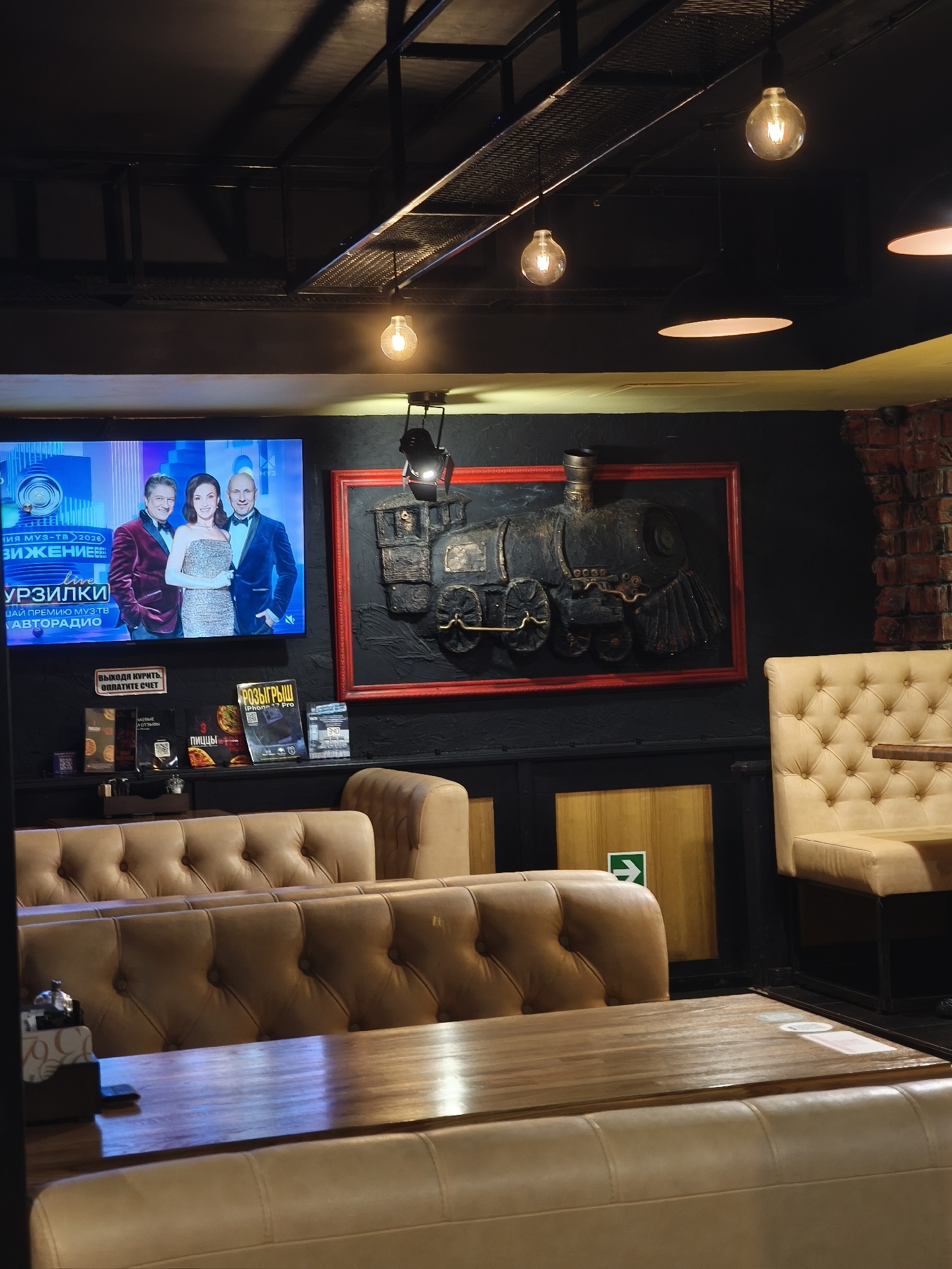 The image captures the interior of a restaurant or bar, likely in the evening, with a dark, industrial aesthetic. The ceiling features exposed metalwork and various light fixtures, including Edison-style bulbs and spotlights, casting warm light on the scene.
A prominent flat-screen television on the left wall displays what appears to be a live awards show, indicated by the "live" text and "МУЗ-ТВ 2026" (Muz-TV 2026) in Russian. Three well-dressed individuals – two men in suits and a woman in a formal dress – are posing on the screen. Below the main text, words like "ЗИЖЕНИЕЕ", "УРЗИЛКИ", "ЦАЙ ПРЕМИЮ МУЗ-ТВ", and "АВТОРАДИО" are visible, further suggesting a televised event related to music awards and radio.
To the right of the television, a large, dark, three-dimensional artwork of a vintage steam locomotive is displayed within a bright red frame against the dark wall. Below the TV and train artwork, a shelf holds various items, including magazines and signs. One sign in Russian reads: "ВЫХОДЯ КУРИТЬ, ОПЛАТИТЕ СЧЕТ" (When going out to smoke, pay your bill). Another magazine cover prominently advertises a "РОЗЫГРЫШ iPhone 17 Pro" (Giveaway iPhone 17 Pro).
The seating consists of comfortable, light brown or beige tufted leather booths and upholstered chairs, arranged around wooden tables. A small silver bell is visible on one of the tables in the foreground. An emergency exit sign with a white arrow on a green background is also visible on a wooden panel on the wall. A section of brickwork on the far right adds to the rustic, industrial decor. The overall setting suggests a casual yet stylish dining or lounge area.