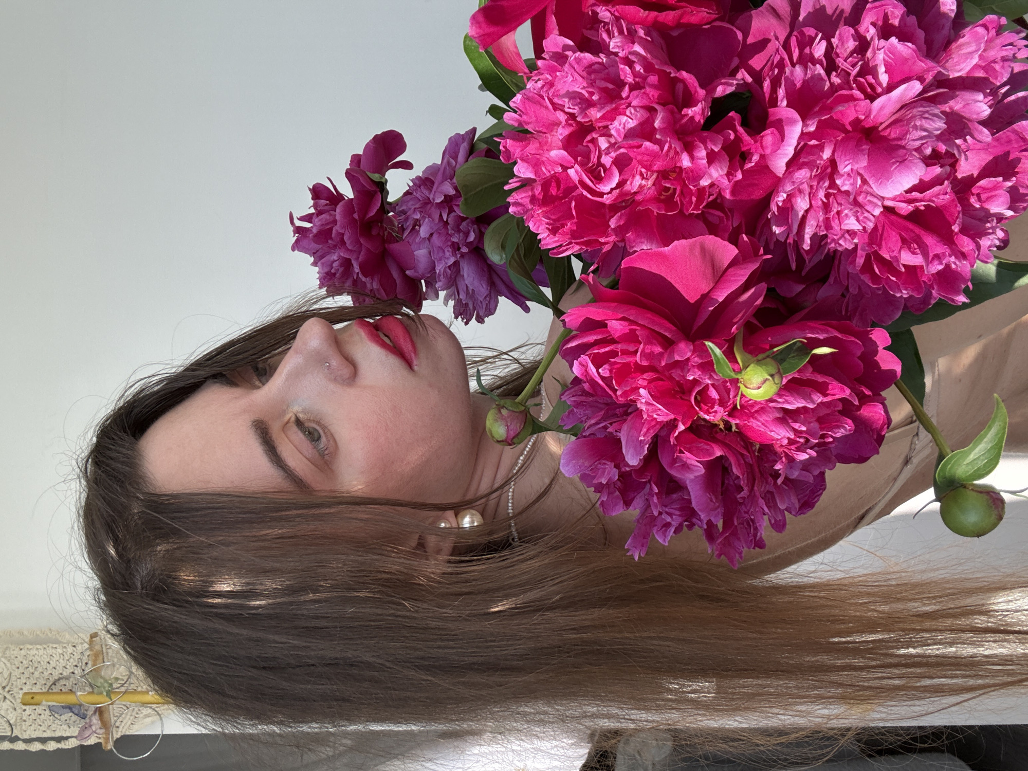 In the foreground, a woman with long, brown hair is shown in profile, her face turned away from the camera. She is wearing bright pink lipstick and a silver nose ring. Her right ear is adorned with a large pearl earring, and a pearl necklace is visible around her neck. She is holding a bouquet of vibrant pink peonies, with several large blooms and a few buds. The flowers are rich in color and texture, with delicate petals. The woman's gaze is directed towards the flowers, and her head is slightly tilted as if she is smelling them.
Behind her, the background consists of a plain white wall. To the left of the woman, a glimpse of a white curtain or sheer fabric is visible, suggesting an indoor setting. A macrame wall hanging with a yellow rod is also present in the upper left corner, adding a touch of texture to the scene. The lighting is natural and warm, casting soft shadows and highlighting the details of the flowers and the woman's features. The image appears to be taken in a well-lit room, possibly a studio or a home.