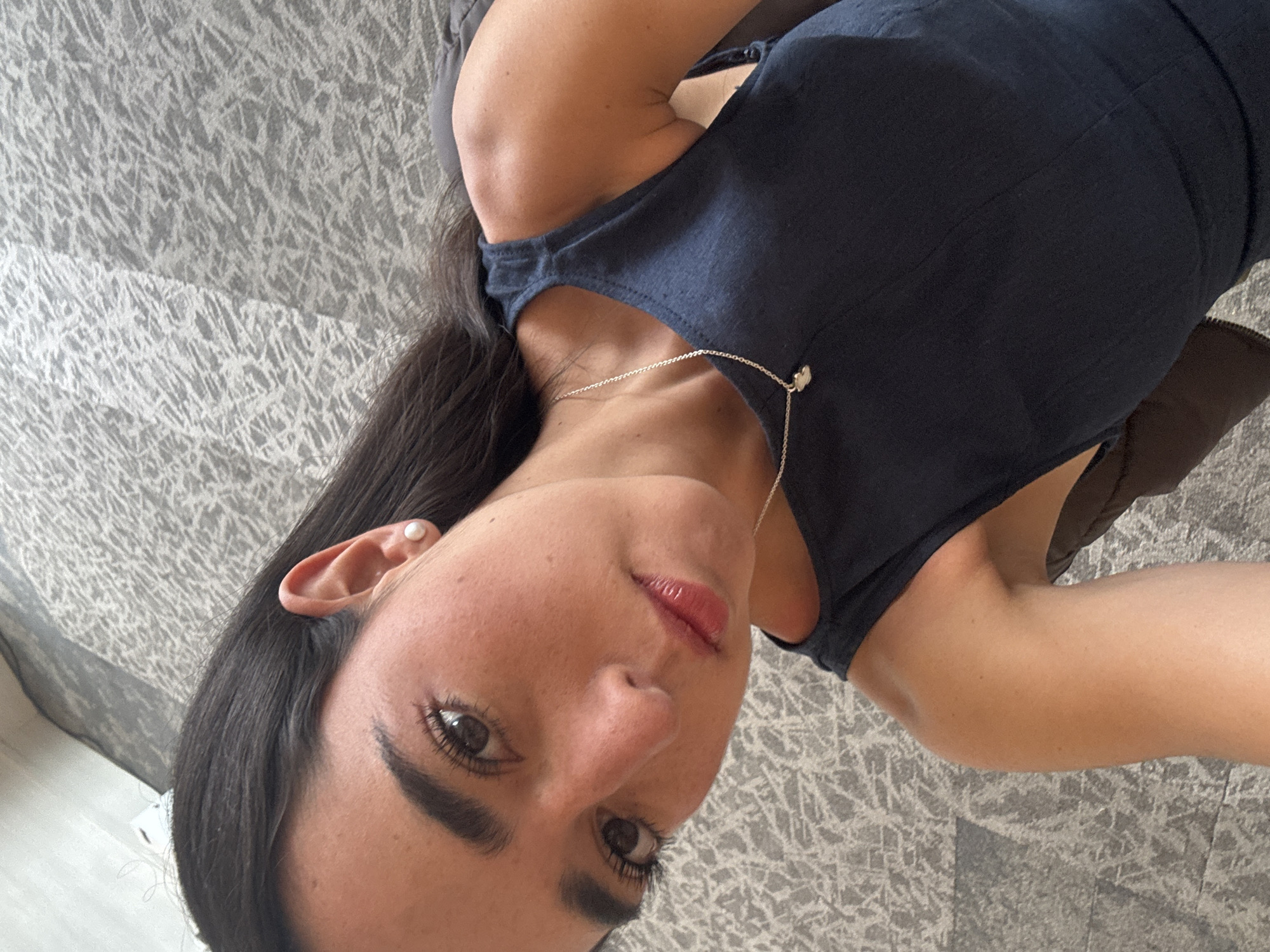 A woman with long dark hair is lying on a gray patterned surface. She is wearing a navy blue dress and a silver necklace with a small charm. A pearl earring is visible in her left ear.