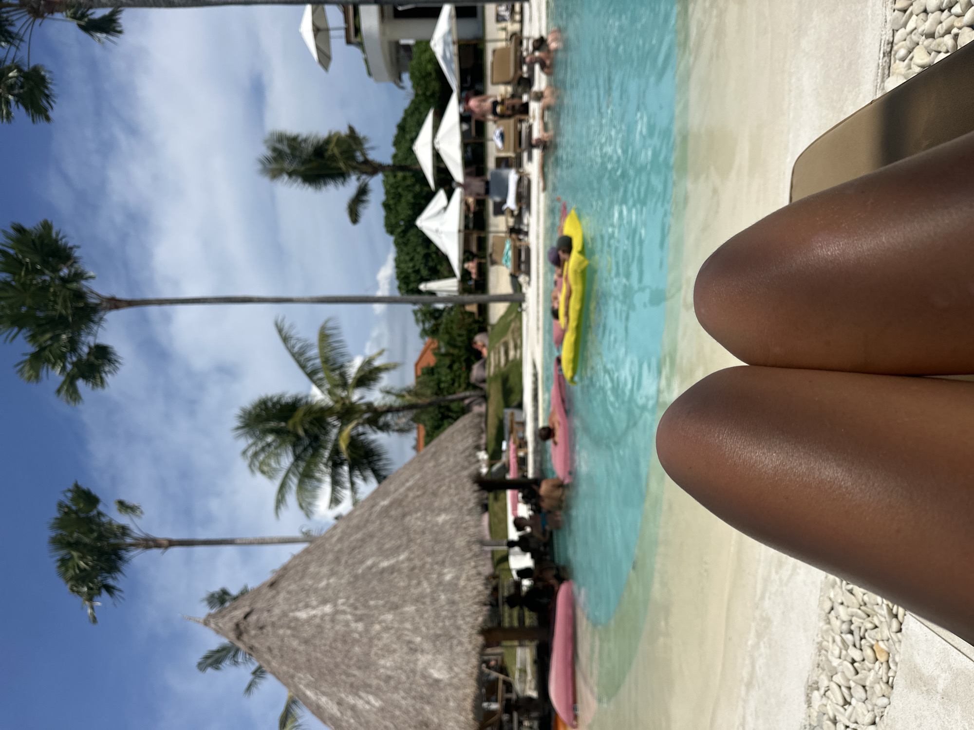 The image shows a sunny scene at a resort-style pool in what appears to be a tropical location. Palm trees are visible against a blue sky with scattered clouds. In the background are white umbrellas and a building. The pool has several people in it, some on colorful inflatable floats. In the foreground are the thighs of a person sitting at the edge of the pool. A thatched roof structure is also visible in the lower left. The location is likely in North Kuta, Indonesia, given the tropical vegetation and resort setting.