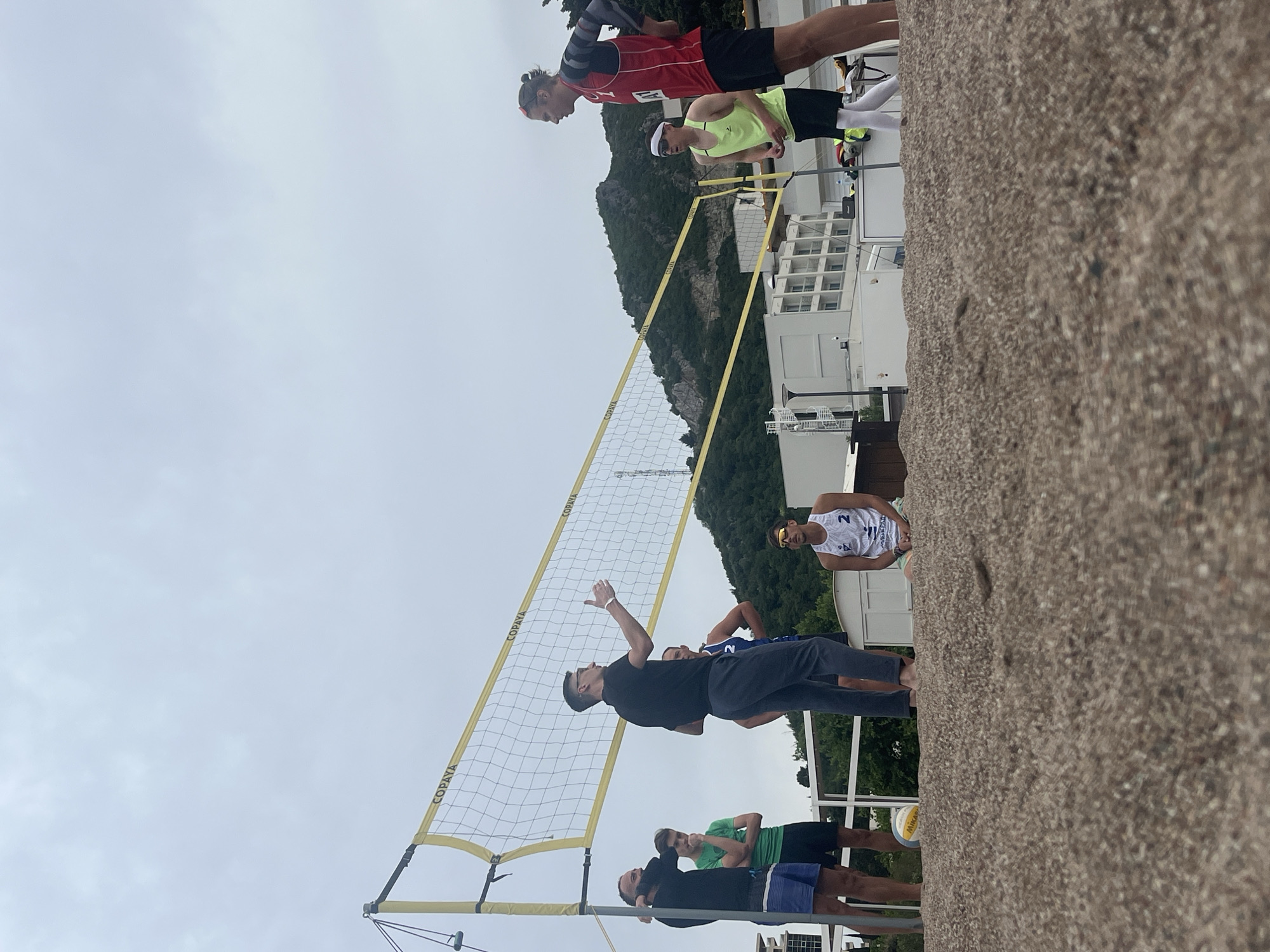 A volleyball net is oriented vertically in front of a group of men on a beach in Boreti, Montenegro. The net has “COPAYA” printed on the yellow support straps. One man wearing a dark-colored shirt and pants is reaching his left arm upward, likely in the act of hitting a volleyball. Other men stand nearby, some looking toward the net and the man reaching. In the background, there are buildings, a mountain partially covered with green vegetation, and a grey sky. A concrete structure is at the bottom right of the frame.