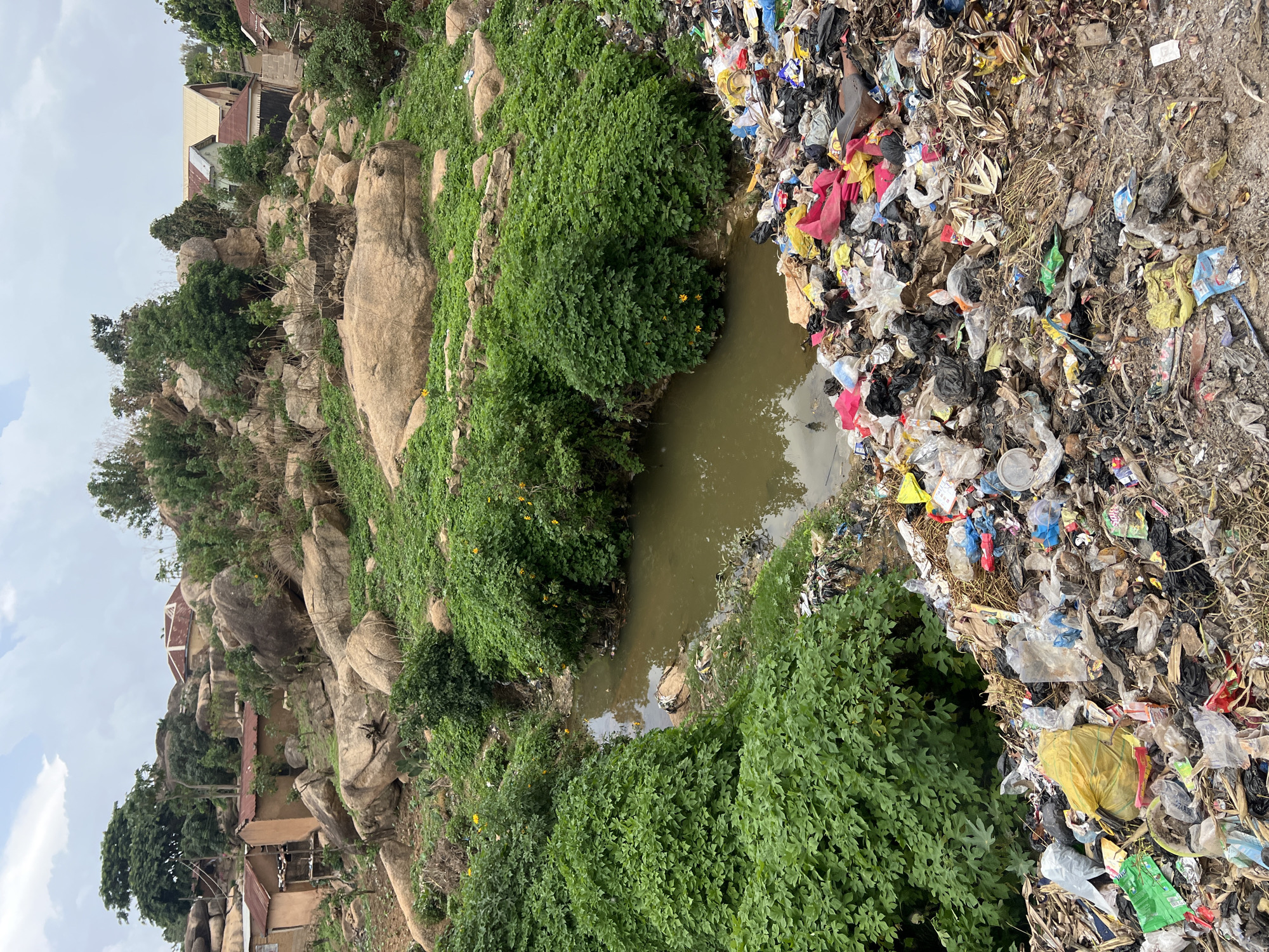 The image depicts a polluted urban area in Jos, Nigeria. A body of water, possibly a stream or pond, runs through the scene, bordered on one side by natural rock formations and dense green vegetation. The opposite bank is covered in a large pile of garbage, including plastic bags, food containers, and general refuse. In the background, simple buildings and dwellings are situated atop the rock formations, indicating a populated residential area. The sky is overcast.