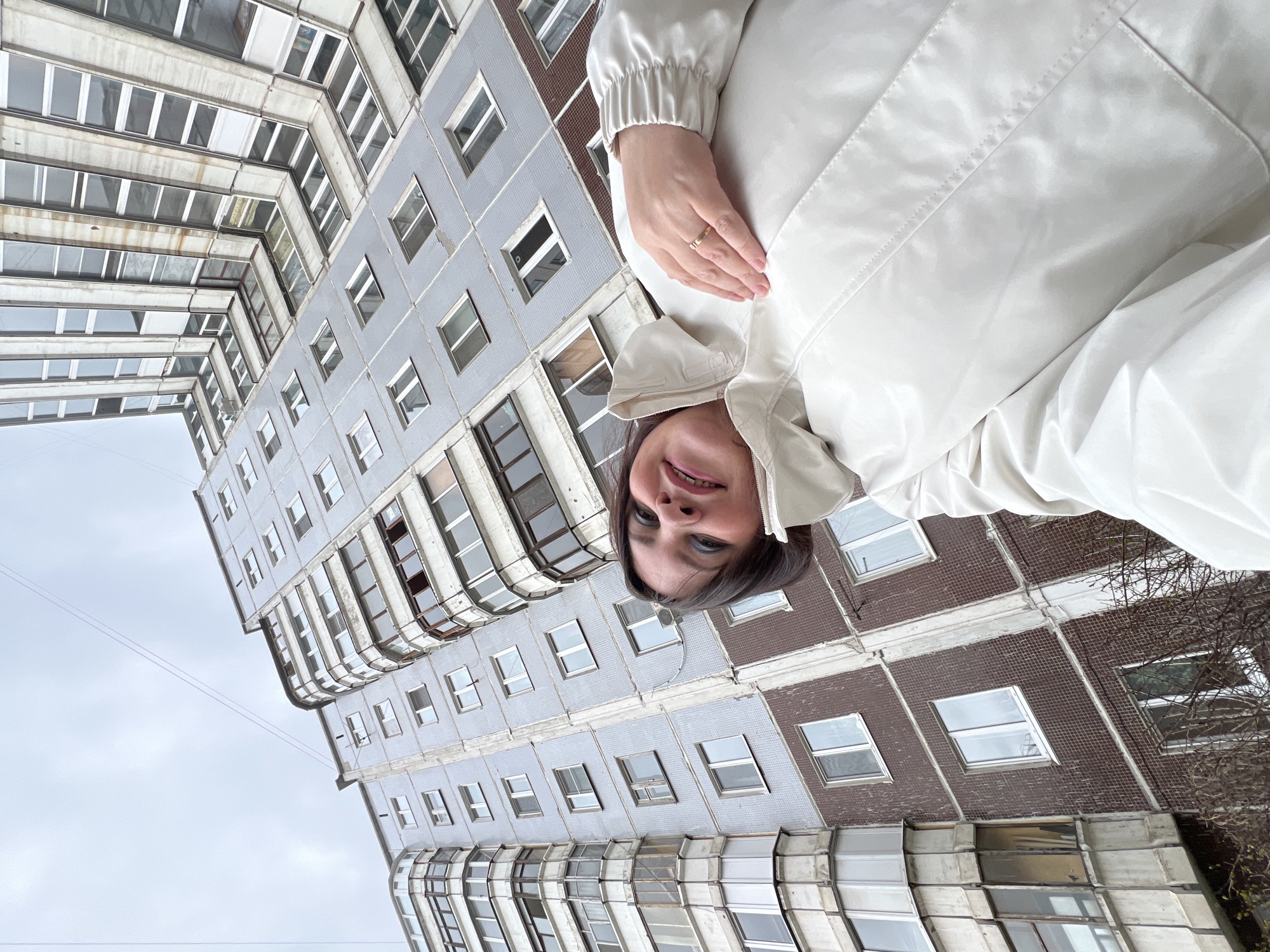 An upward-angled, rotated photograph captures a woman smiling against the backdrop of a multi-story apartment building in Perm, Russia, under an overcast sky. The image is rotated counter-clockwise, with the building spanning from the lower left to the upper right, and the woman positioned in the lower right foreground.
The primary subject is a woman with dark hair and a light complexion, looking directly at the camera with a wide, open-mouthed smile, revealing braces on her lower teeth. She is wearing a light, possibly off-white or cream-colored, shiny jacket with a high collar. Her right hand, adorned with a simple band ring on her ring finger, rests on the jacket over her midsection.
The background features a large, angular residential building characterized by its repeating pattern of windows and a facade primarily composed of light blue/grey and reddish-brown square tiles. The building exhibits architectural elements like curved bay windows and recessed sections, suggesting a functional, possibly Soviet-era design. Several windows are visible, some appearing slightly open. The sky is uniformly light grey, indicating an overcast daytime. Bare tree branches are visible near parts of the building. Two thin power lines stretch across the sky in the middle distance. No visible text is present in the image.