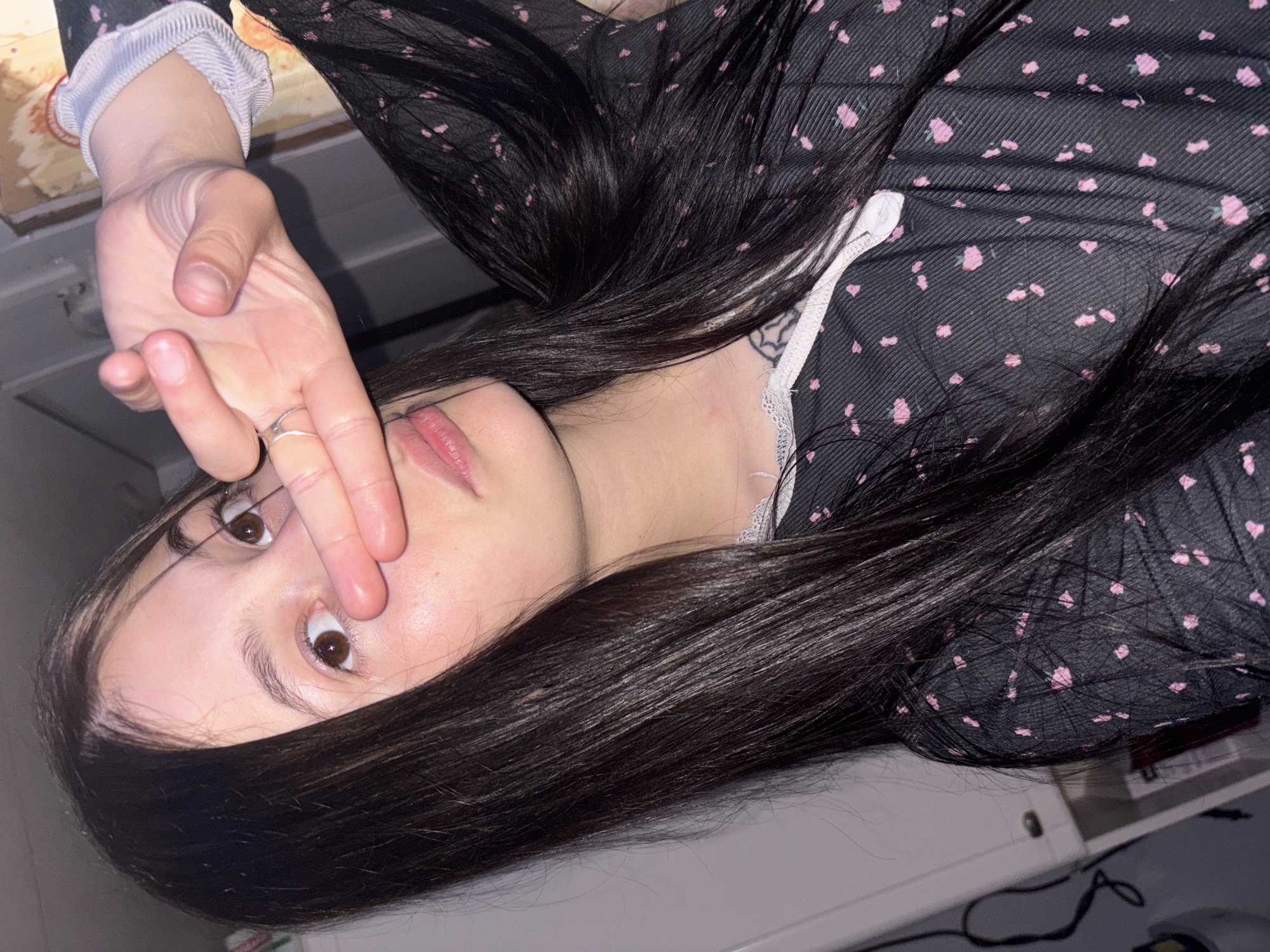 A close-up portrait of a young person, likely female, is captured indoors, possibly in a room in Фоки, Russia. The subject has long, dark, straight hair cascading over her shoulders and chest. She is dressed in a dark garment, appearing to be a dress or top, patterned with small pink floral designs, and features white lace trim around the neckline. A light-colored, subtly striped sleeve is visible on her left arm, adorned with a silver ring on her left ring finger.
Her left hand is raised to her face, with fingers gently covering her nose and upper lip, creating a gesture that conveys shyness, playfulness, or contemplation. Her brown eyes are open and directed towards the viewer. A subtle tattoo can be glimpsed on her neck/shoulder area, partially hidden by her hair and clothing.
The background shows parts of a light-colored surface, possibly a desk or table, along with a dark cable and another indistinct object. The lighting suggests an indoor setting, likely under artificial light, possibly in the evening or at night. No text is visible in the image.