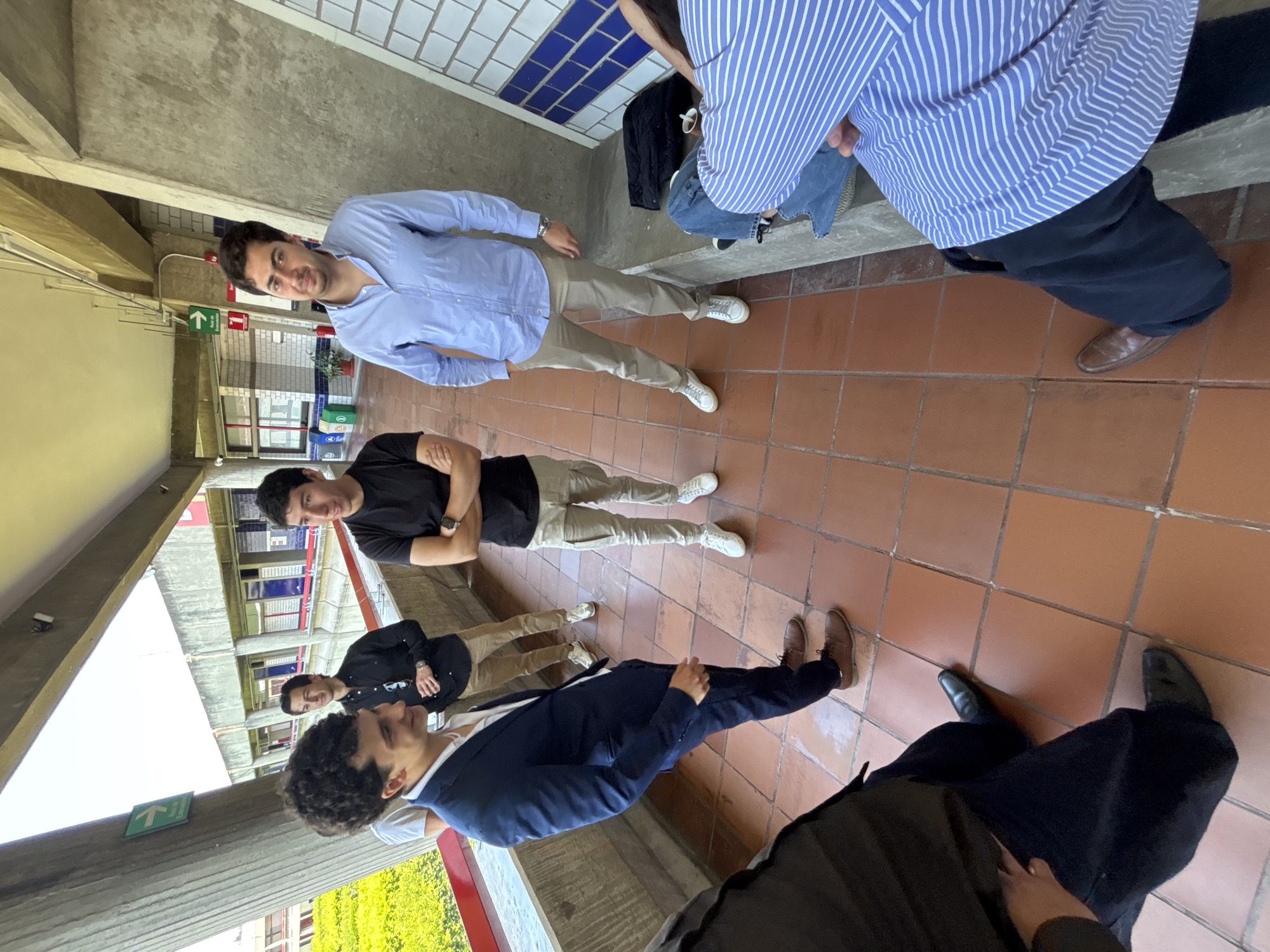A group of five men are gathered indoors, likely in a building with architectural elements typical of the city of Puebla, Mexico. The floor is tiled with red-brown squares. Three of the men are standing, and their full bodies are visible. The other two are partially visible. One man wears a light blue button-down shirt and khaki pants with white sneakers. Another wears a black t-shirt, khaki pants and white sneakers. The third man visible is wearing a dark blue suit jacket with brown shoes. The partially visible men are wearing a striped blue and white shirt and dark trousers with brown shoes, and the other is wearing a black shirt and khaki pants. Overhead, there are signs with a green arrow indicating an exit. The background shows a multi-level building with visible windows and possibly green vegetation. A bag is sitting against a wall with white and blue tiles.