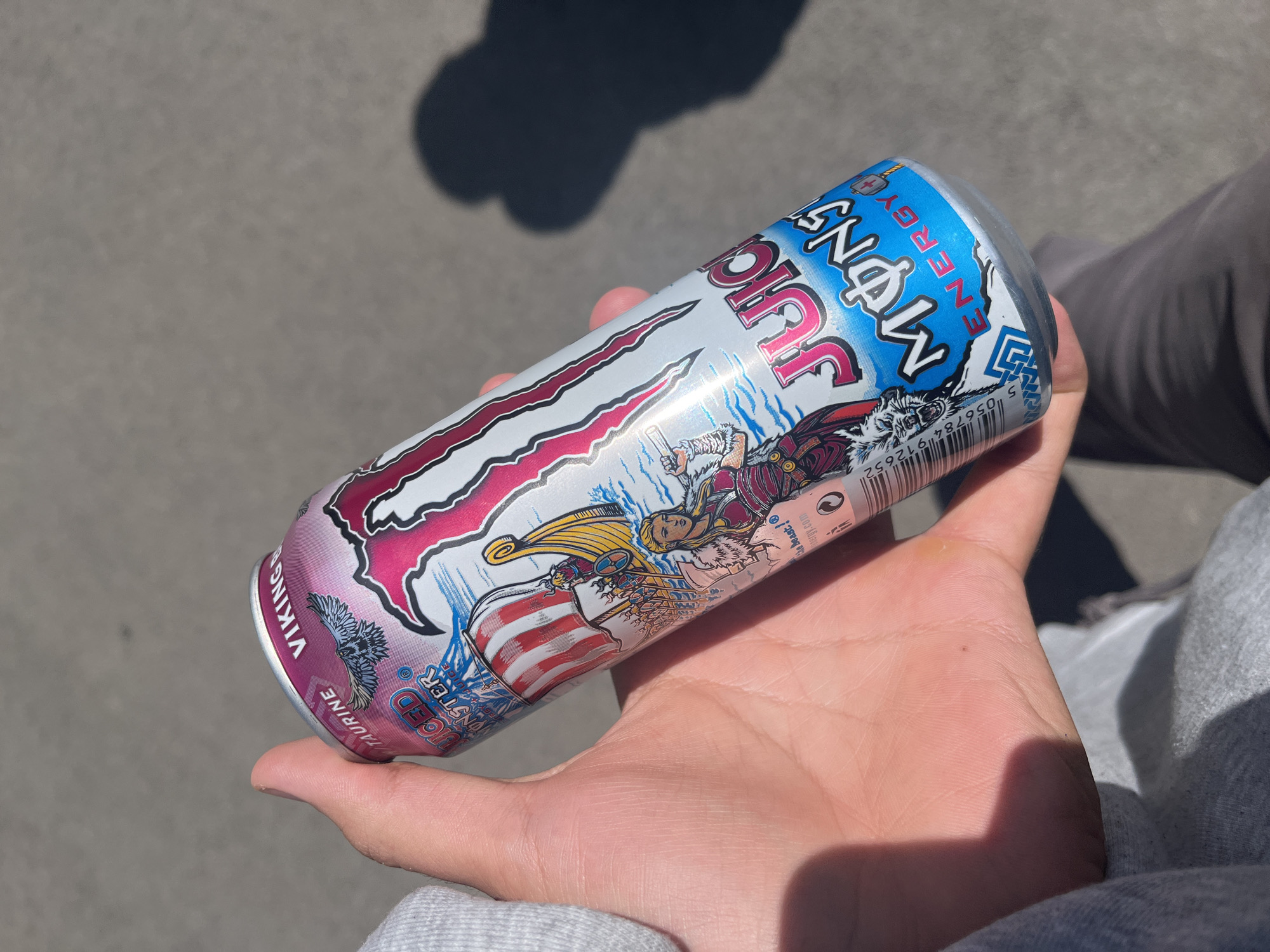 A person's hand holds a Monster Energy drink can in an outdoor setting in Sofia, Bulgaria. The can is tilted, showcasing its colorful design. The ground beneath is a light grey paved surface, likely concrete or asphalt, with strong shadows suggesting bright sunlight.
The Monster Energy can is predominantly white with a large pink "M" logo, characteristic of the brand. Text on the can includes "JUICE MONSTER ENERGY" in blue and pink, and "VIKING" printed vertically, indicating the specific flavor or variant. Detailed illustrations of Viking-like characters, including one with a horned helmet and another with a longboat, are visible on the can's label. Additional text such as "TAURINE" and "VICED MONSTER" can be seen near the bottom. A partial barcode and "the beast! ergy.com" are also discernible.
The person holding the can is wearing a grey hooded garment on their left arm, and dark pants are visible in the background on the right. The strong shadows on the ground indicate a sunny day. The scene captures a casual moment of someone holding a beverage outdoors.