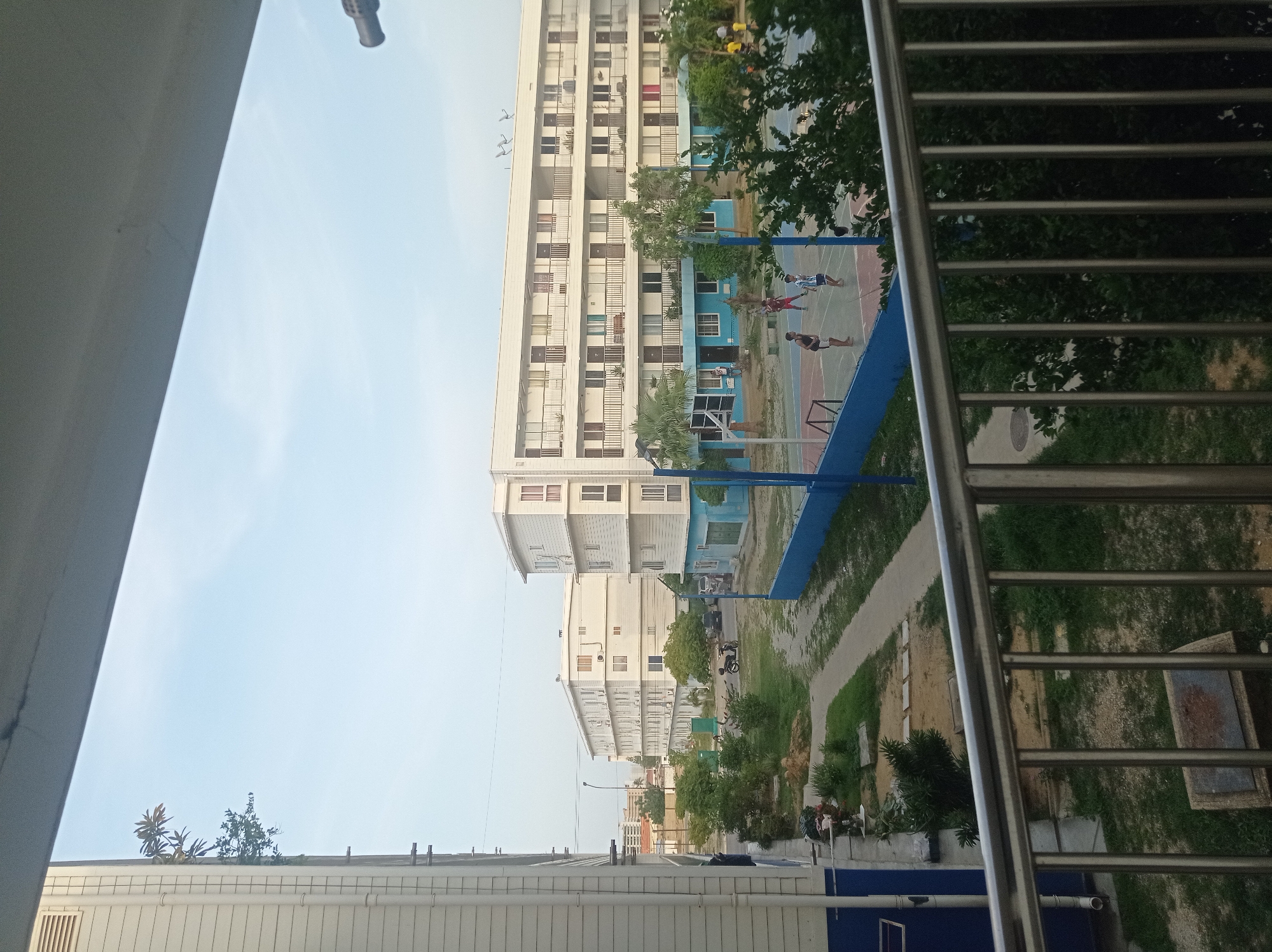 The image captures a view of a residential complex in Porlamar, Venezuela, seen from a balcony with a metal railing in the foreground. The complex features several multi-story white buildings with blue trim, many featuring balconies with dark railings. A paved courtyard area with a basketball court, where several individuals are engaged in a game, is visible between the buildings. There are also trees and green spaces throughout the complex, and a motorbike and car are parked in the courtyard. The sky is clear and pale blue, suggesting it is daytime. The scene conveys a sense of everyday community life within the urban environment.