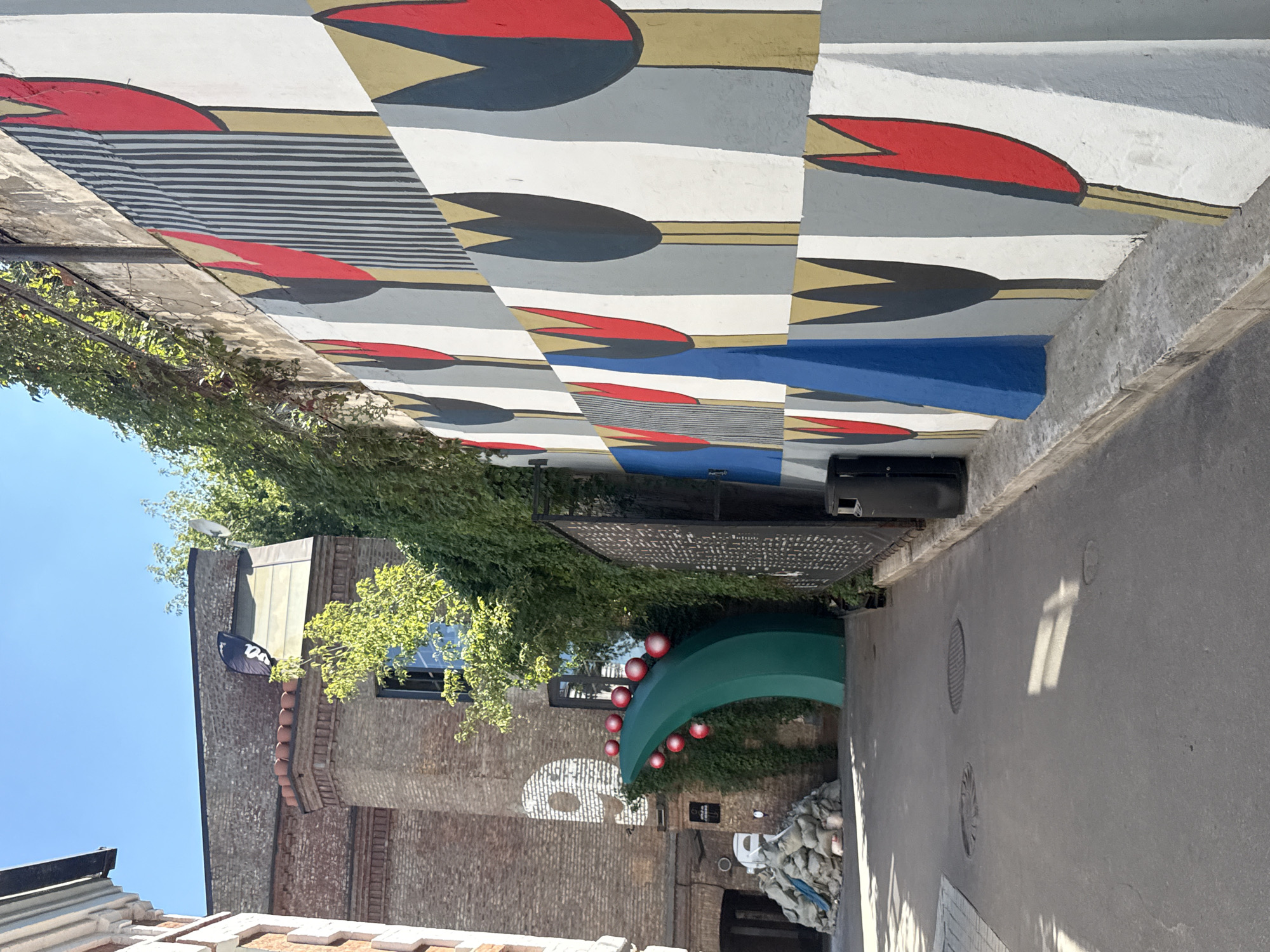 The image captures an outdoor urban scene, likely a courtyard or alleyway, in Moscow, Russia. The focal point is a vibrant mural covering a long wall on the right side of the frame. The mural depicts stylized tulips in red, gray, and yellow, set against a background of white and vertical black stripes. The design is geometric and repetitive, creating a dynamic visual effect.
To the left of the mural, a building with a brick facade is partially visible. Ivy and other greenery cascade down from a higher level, softening the urban architecture. In the foreground, a paved pathway runs between the muraled wall and the building. Several manhole covers are embedded in the pathway.
Further down the pathway, a large, curved green structure is installed, adorned with several red spherical objects. Behind this structure, a white spray-painted symbol or logo is visible on the brick wall, resembling the number "9" with a stylized face. A pile of sandbags is situated to the left of the green structure. A black trash receptacle stands against the muraled wall near the center of the frame.
The sky above is clear and blue, suggesting a sunny day. The overall impression is one of a creatively decorated urban space, blending art, architecture, and nature.
