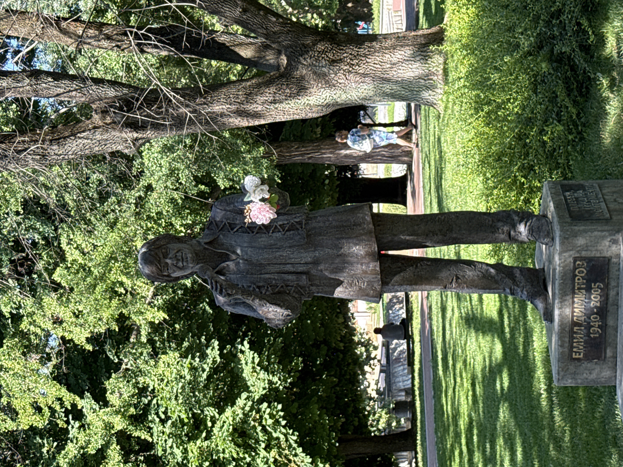 The image shows a bronze statue of a man holding a bouquet of flowers. The statue is located in a park with lush green trees and grass. In the background, a woman is walking, and a person is sitting on a bench. The base of the statue has an inscription that reads "ЕМИЛ ДИМИТРОВ 1940-2005", identifying the subject as Emil Dimitrov, a Bulgarian singer. The statue is a monument in Pleven, Bulgaria.