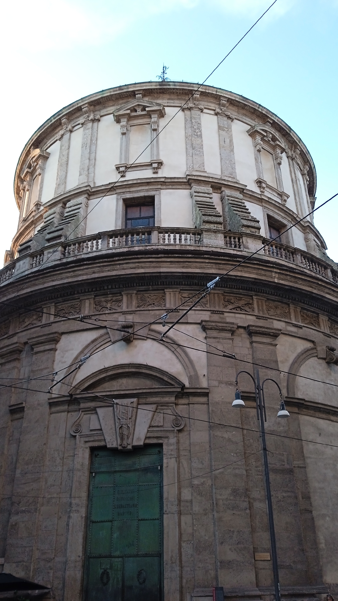 The image shows a low-angle view of the circular facade of San Sebastiano church, located in Milan, Italy. The church is made of stone and features a dome and ornate architectural details. The facade has a large green door with inscriptions, a balcony with balusters, and several windows. Overhead, electrical wires crisscross the sky, and a lamppost with two lights is visible on the right side of the image. The sky is light blue and mostly clear. There are no people visible in the frame.