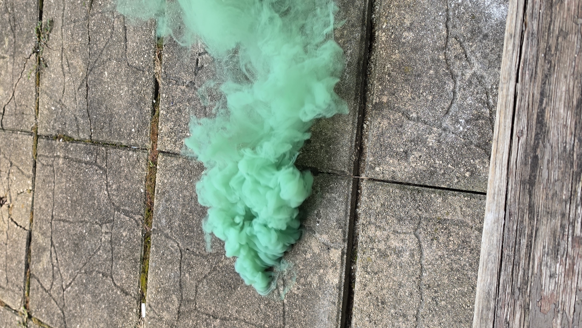 The image shows a plume of bright green smoke emanating from the left side and dispersing to the right. The smoke is dense and appears to be a vivid, almost artificial, green color. It is situated on a surface composed of large, grey concrete pavers. These pavers have visible cracks and some patches of moss or dirt in the crevices between them, suggesting an outdoor environment. In the foreground, a weathered wooden plank is visible, running horizontally across the bottom of the frame, with visible grain and some peeling paint. The scene appears to be outdoors on a paved area, possibly a patio or walkway. There are no people, animals, or other discernable objects present besides the smoke, pavers, and wood. The image does not provide any information about the location within the city of Elgin, United States of America.