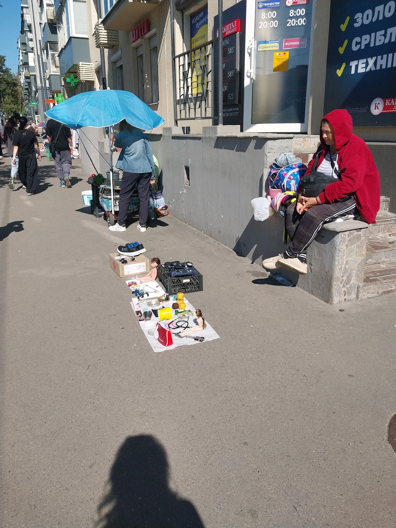 The image depicts a street scene in Dnipro, Ukraine, on a sunny day. In the foreground, a person wearing a red hooded sweatshirt is sitting on concrete steps, facing left and looking down. Next to them are several bags and a backpack. To the left of the seated person, on the sidewalk, a collection of items is laid out on a white cloth, including toys, a red bag, and various small objects. Behind this display, a blue umbrella is propped up, providing shade. Several people are walking on the sidewalk in the background, and a cart is visible near the umbrella. The environment includes a building with storefronts, one of which has an advertisement with text in Ukrainian. The pavement is asphalt, and shadows are cast on the ground.