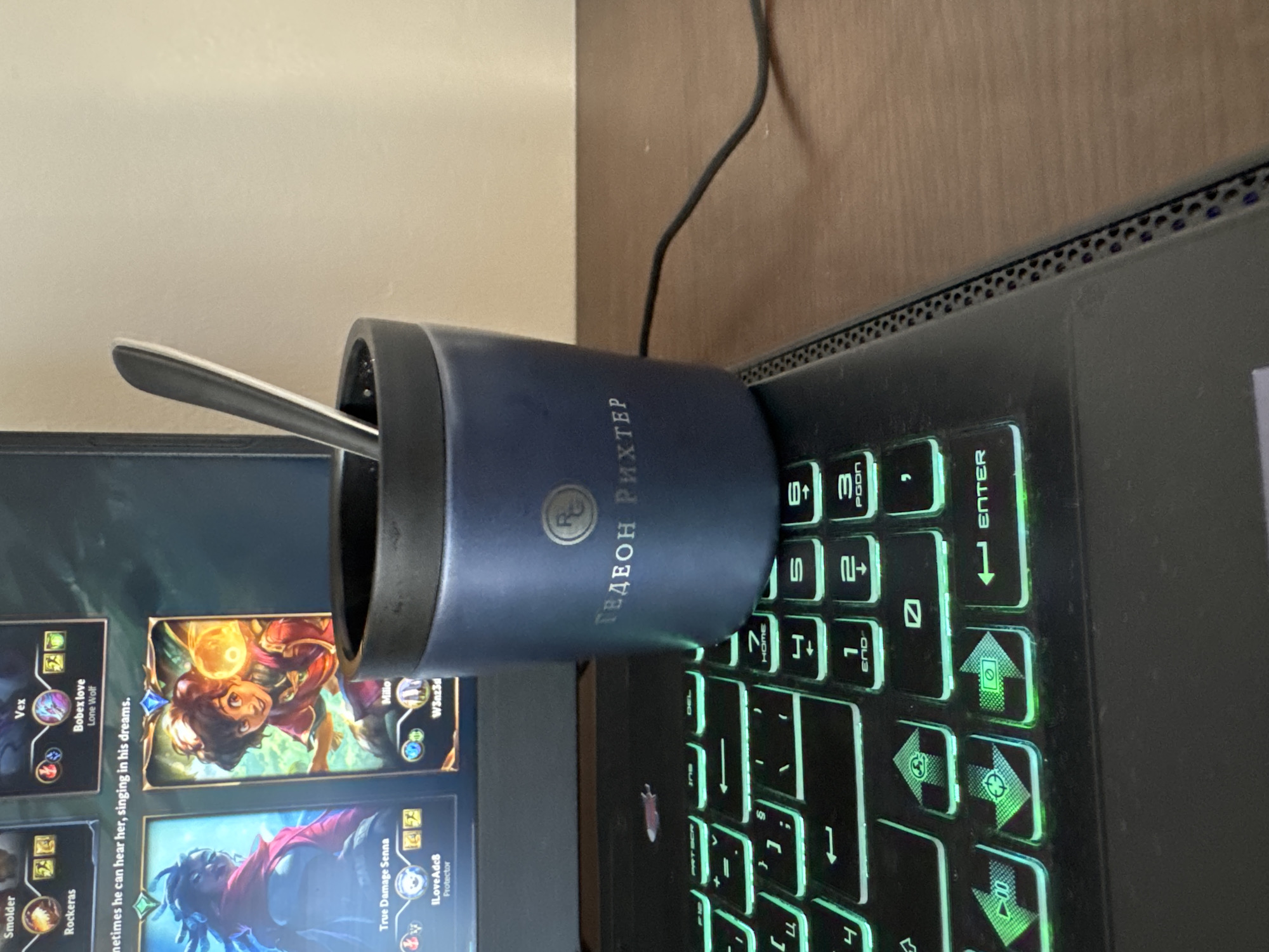 The image depicts a desk with a laptop running a card game, likely Legends of Runeterra. A blue cup, branded with "ГЕДЕОН РИХТЕР" and an emblem, sits on the right side of the laptop near the keyboard. A spoon is inside the cup. The laptop keyboard is illuminated with green backlighting. There is a cable extending from the top right of the laptop. The background is a plain wall. The image was taken indoors. The laptop screen displays various game cards, including characters named Smolder, Vex, Milio, and True Damage Senna. The location is unknown and cannot be determined from the visual information provided.
