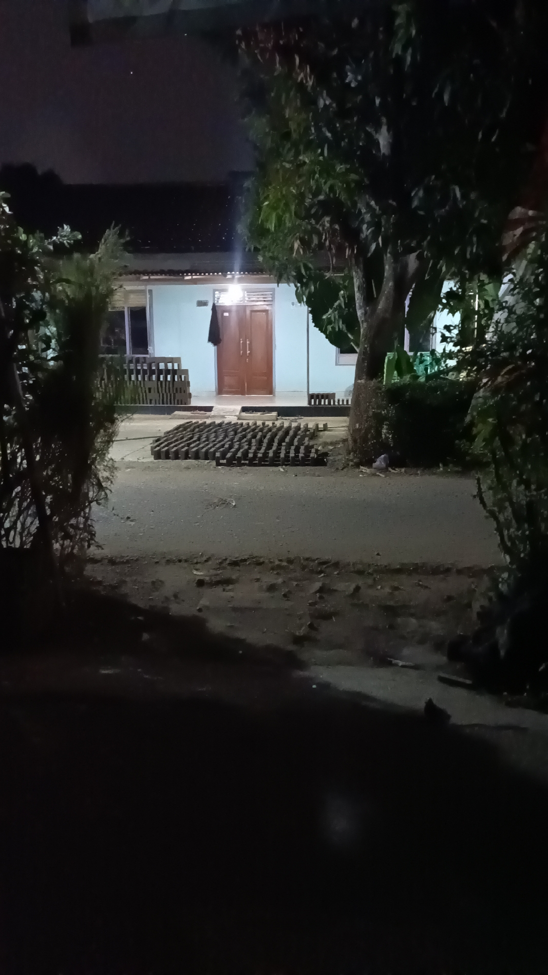 The image depicts a nighttime scene outside a light blue building with double wooden doors. A dark garment hangs on the right door. Rows of bricks are stacked on the left side of the building, and a larger rectangular arrangement of bricks lies on the asphalt road in front of the building. Two trees are prominent, one to the right of the building and another to the left, partially obscuring the view. The sky is dark with a hint of clouds. The foreground is very dark. The location is described as being in the city of Kebumen, Indonesia.