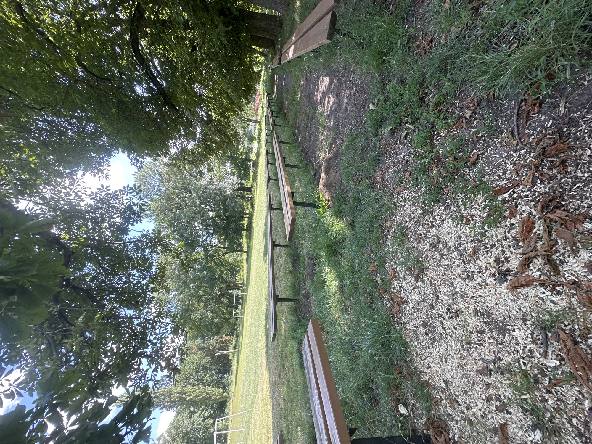 The image depicts an outdoor scene featuring multiple rows of park benches situated on a grassy area with a dirt path winding through. In the background, there are trees and what appears to be a children's playground with a swing set and a soccer goal. The scene is bathed in natural daylight, suggesting it is daytime. The specific location in Makiv, Ukraine, is not visually discernible from the image itself, as there are no identifying landmarks or signage present. The benches are empty, indicating no immediate human presence or activity at the time the photograph was taken. The environment suggests a park or recreational area, possibly a public space designed for relaxation or outdoor activities.