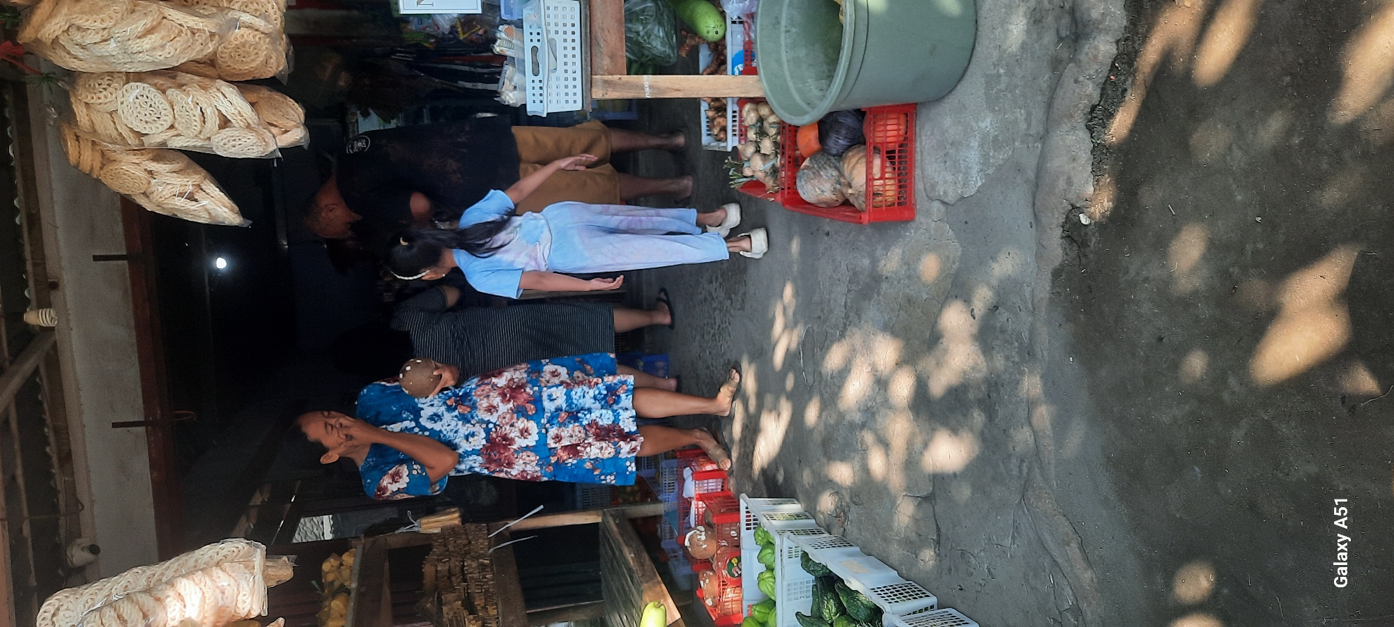 The image depicts a street-side market stall in Selopuro, Indonesia, with several individuals present. A woman in a floral blue dress stands in the foreground, holding a coconut and covering her mouth with her hand, possibly laughing or talking. To her right, a young girl in a light blue tie-dye outfit faces away from the camera, walking towards the market stall. Behind them, two men are visible; one in a black t-shirt and shorts, and another partially obscured by the woman. The stall is laden with various produce, including green gourds in white crates, red crates filled with what appear to be pumpkins and other vegetables, and bags of light-colored crackers hanging above. The ground is paved and shows patches of sunlight and shadow, suggesting an outdoor setting. The overall scene captures a moment of daily commerce in the Indonesian locale.