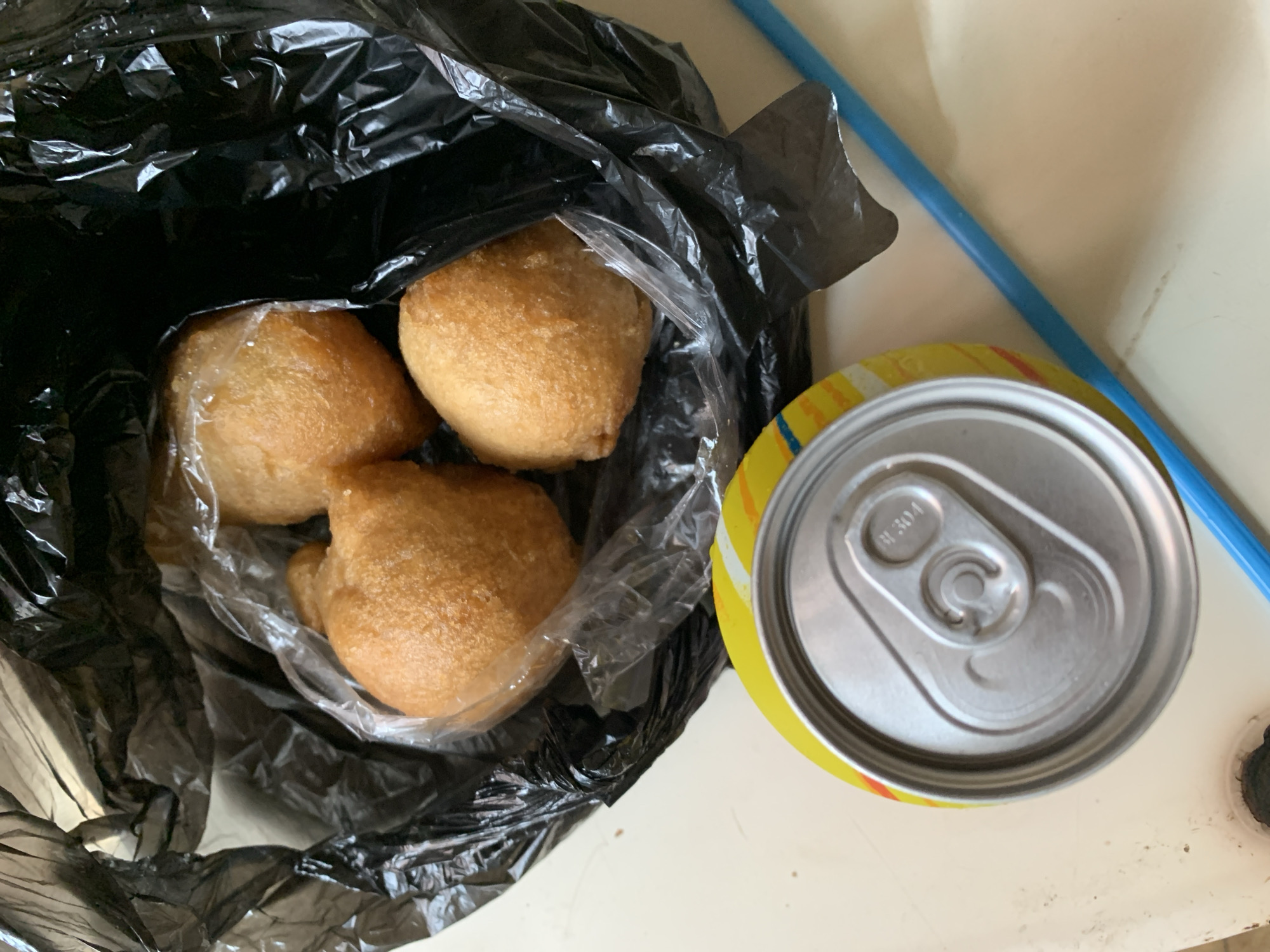 In this top-down view, three golden-brown fried snacks are nestled inside a black plastic bag. The snacks, which appear to be a type of fritter or pastry, are partially wrapped in clear plastic. To the right of the bag sits a silver soda can with a yellow and red label, its pull-tab clearly visible. A thin blue object, possibly a straw or a pen, runs diagonally across the background. The scene is set on a light-colored surface, likely a table or countertop, with subtle hints of dirt or crumbs. The lighting suggests an indoor setting, possibly during daylight hours, given the brightness. The overall presentation is casual, hinting at a simple snack or meal. No people are visible. The only visible text is on the soda can, which reads "R 304". The context provided places this scene in Kabong, Nigeria.