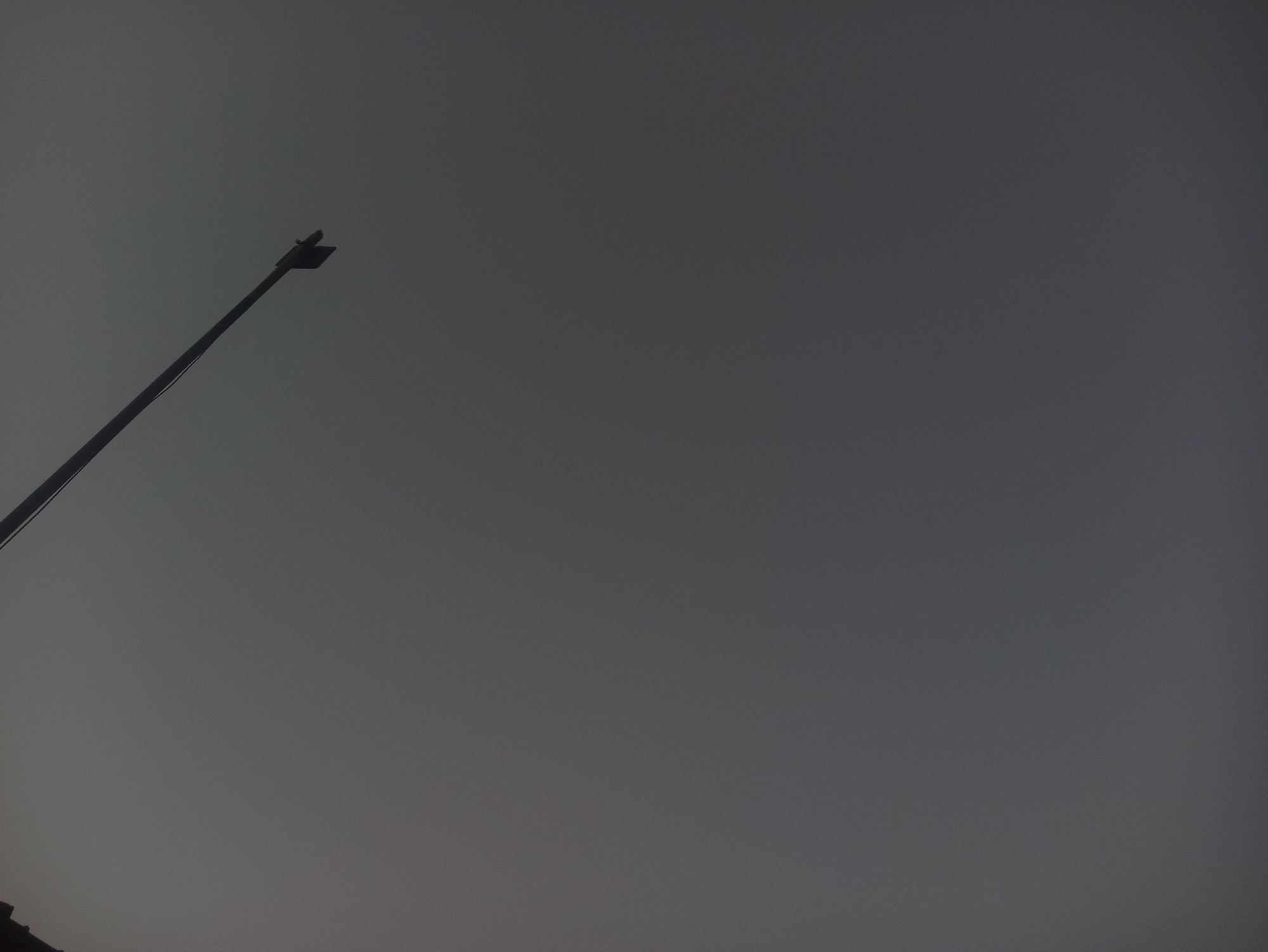 The image shows a silhouette of a street lamp against a dark, overcast sky. The street lamp is positioned on the left side of the frame, extending diagonally upwards from the bottom left corner towards the top right. The sky is a uniform, muted gray, suggesting either twilight or a very cloudy day. There is a hint of dark ground or structures at the very bottom left of the image, but they are largely obscured by the darkness and the focus on the sky and lamp. The scene appears to be an outdoor environment, possibly a street or park, in the city of Tuba, Indonesia, given the context.