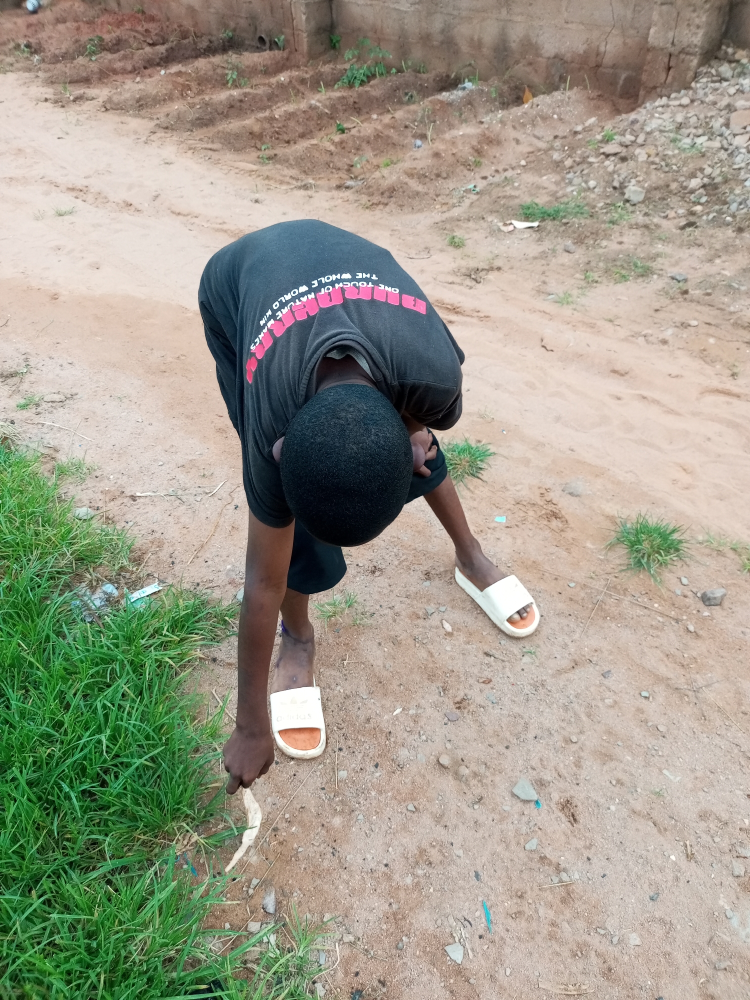 A young boy is bending over to pick something up from the ground. He is wearing a black t-shirt with pink lettering and black shorts. He is also wearing white flip-flops. The ground is a mix of dirt and sand, with patches of green grass. In the background, there is a wall made of concrete blocks. The scene appears to be an outdoor area in a dusty, possibly rural, setting. The location is identified as Tempure, Nigeria.