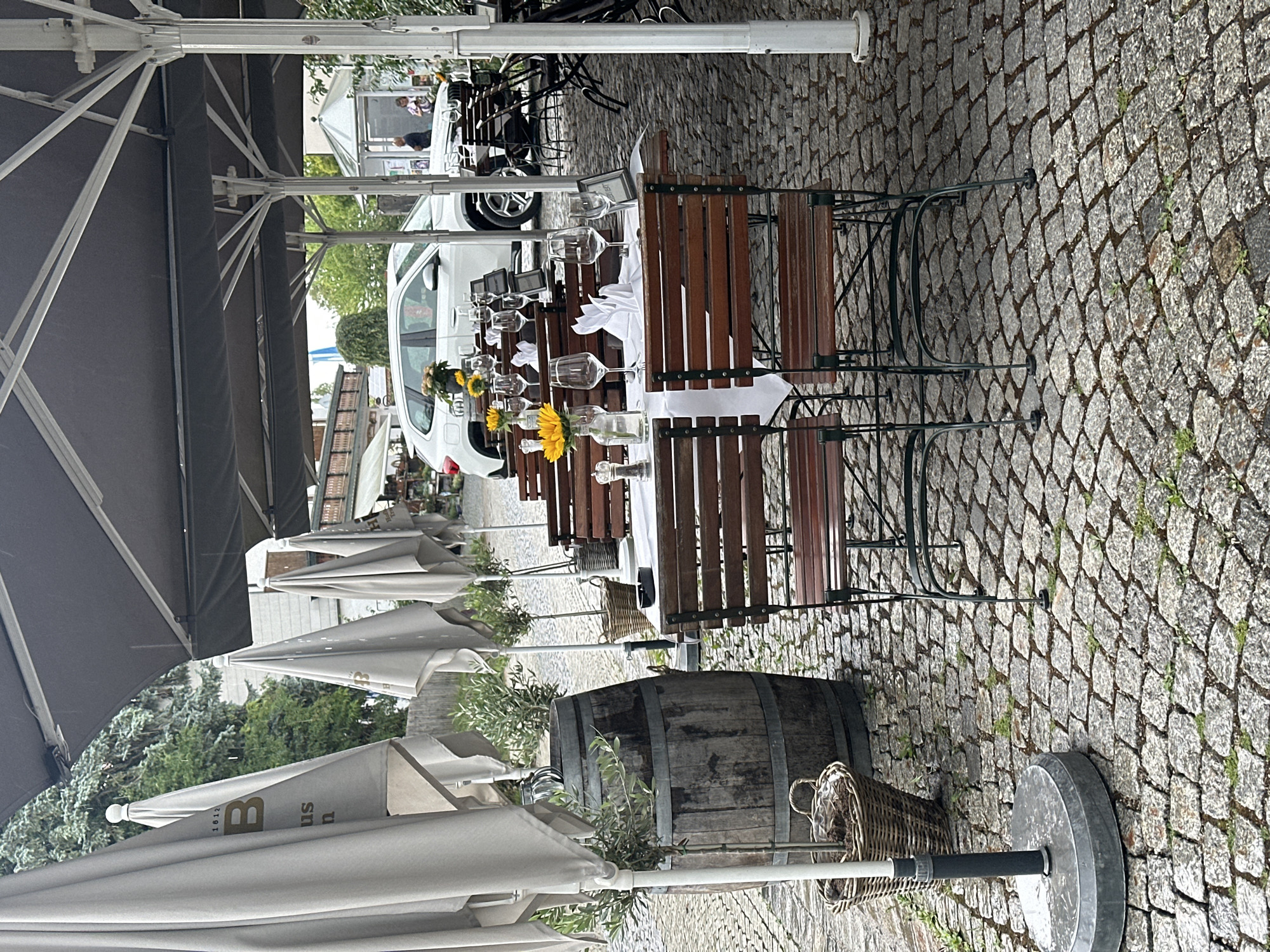 The image depicts an outdoor seating area of a restaurant or cafe in Seeon-Seebruck, Germany, likely during daytime. The scene is set on a cobblestone ground, covered by large grey umbrellas. Several tables are arranged under the umbrellas, set with white tablecloths, wine glasses, and napkins folded into swan shapes. A vase with sunflowers sits on one of the tables, adding a touch of color. In the background, a white car is parked, and a few people are visible near a building. To the left, a large wooden barrel and wicker baskets are placed near a row of closed umbrellas. The overall atmosphere suggests a quiet, possibly rainy, day, as indicated by the closed umbrellas and the damp appearance of the cobblestones.
