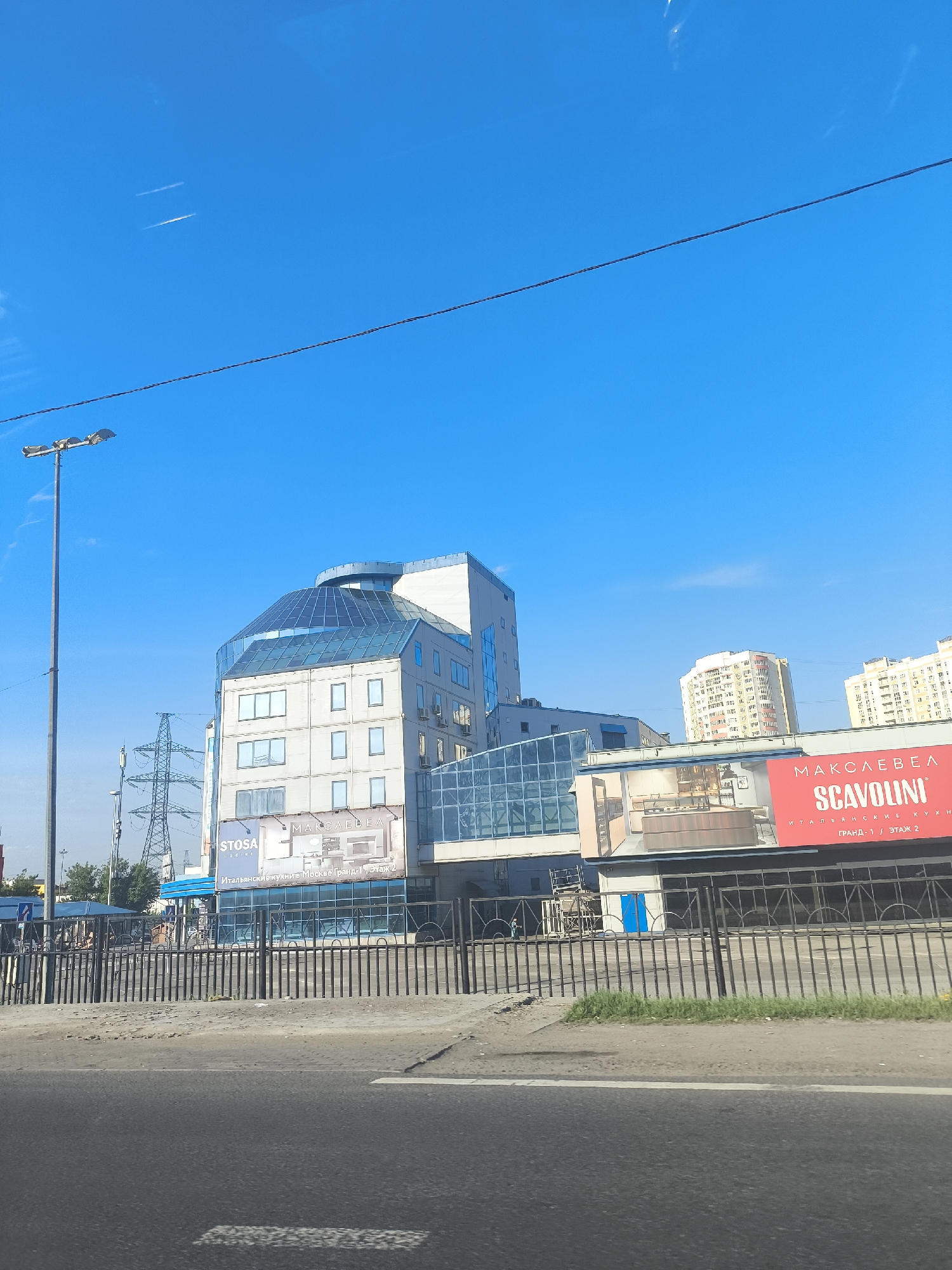 The photograph depicts a street scene in an urban area, possibly the city of Unknown, Unknown. A multi-story building with a modern design, featuring a glass-domed roof and a glass-enclosed walkway, is prominent. A large sign advertising "Scavolini" and "Stosa Cucine" Italian kitchens is visible on the building's facade. A black metal fence runs along the street, separating the sidewalk from the road. A light pole stands tall to the left of the building. Power lines are visible in the background. A pedestrian bus station is visible in the distance. In the far background, apartment buildings rise above the scene. The sky is clear and blue, with a few contrails from aircraft. The photograph appears to have been taken from a moving vehicle on the road.