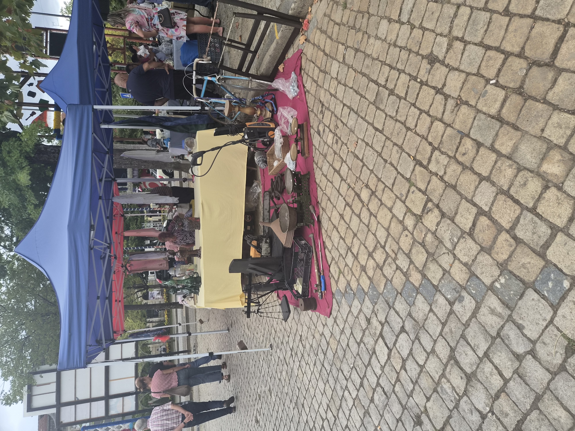 An outdoor market is set up under a large blue canopy tent and a smaller red and pink tent. The ground is paved with cobblestones. In the foreground, a red cloth displays various items, including scales, metal bowls, a lawn mower, a chainsaw, and other tools. Behind this, a yellow-covered table exhibits decorative items like glass bottles and ceramic figures. Several people are visible browsing the stalls and interacting with vendors. A bicycle is parked near the table. In the background, more tents and market activity can be seen, along with trees and buildings. The scene depicts a bustling market day in Barcelos, Portugal.