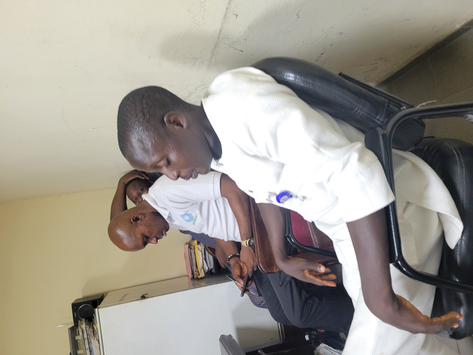 The image captures an indoor scene with three individuals. The primary subject is a man in a white, long-sleeved shirt, seated in a black, leather office chair. A second man, who is bald, is wearing a white T-shirt with a logo on it, and is seated beside him. A third person, whose face is partially obscured, is positioned behind the bald man.
The environment includes a white cabinet with black speakers on top, and some files. It is unclear if the location can be pinpointed to the city of Abuja, Nigeria without additional context. The image does not provide any clues to confirm that the picture was taken specifically in Abuja.