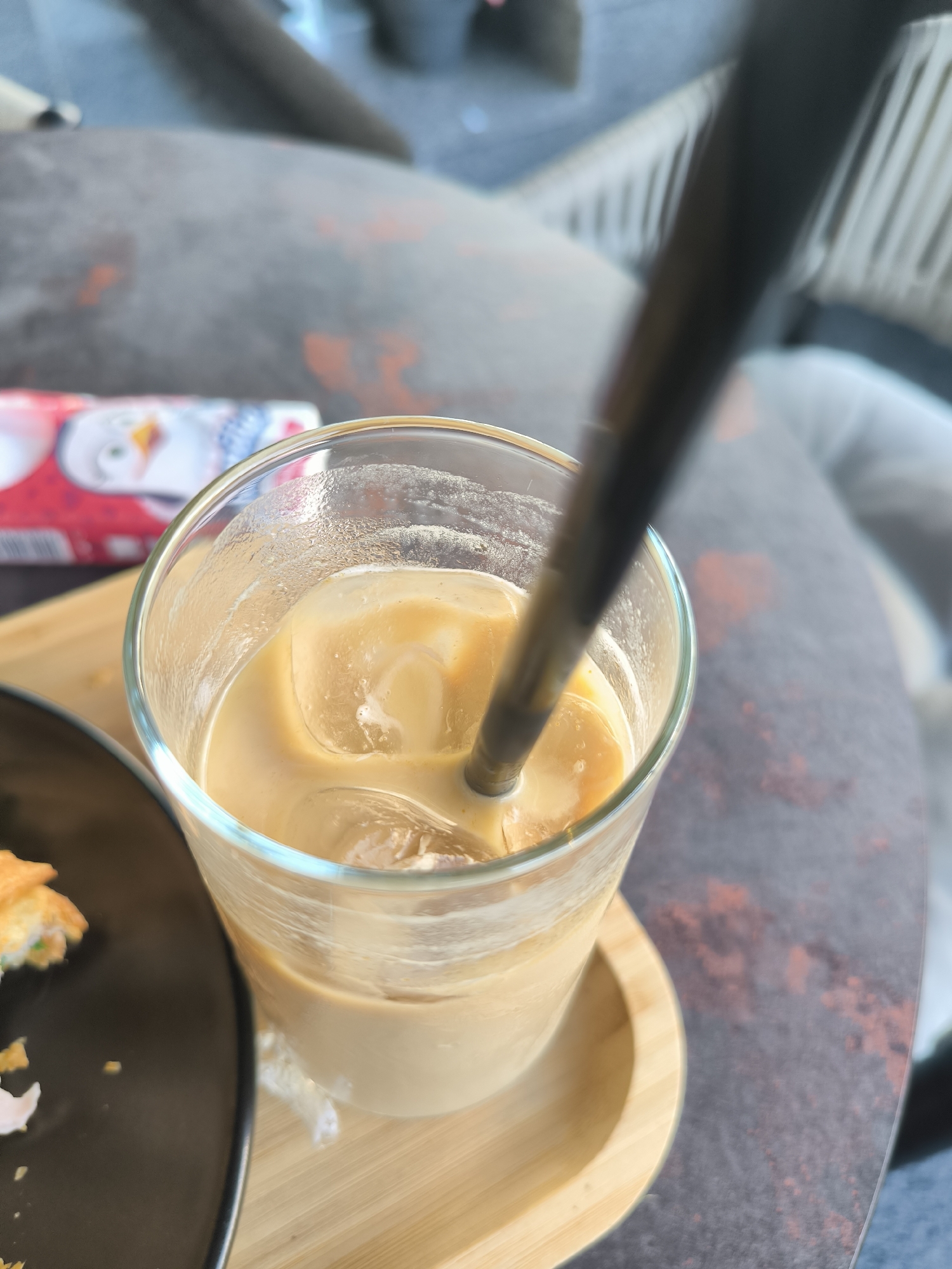 A close-up, overhead shot captures an iced coffee with a black straw in a clear glass, resting on a light wooden coaster. The drink, a light brown color with several ice cubes, is positioned on a dark, textured table. To the left, a portion of a black plate with a pastry is visible. In the background, out of focus, a package with a cartoon penguin design is partially visible. The overall setting appears to be an indoor cafe or restaurant, potentially in Chișinău, Moldova, given the context of the request. There are no identifiable individuals in the frame.