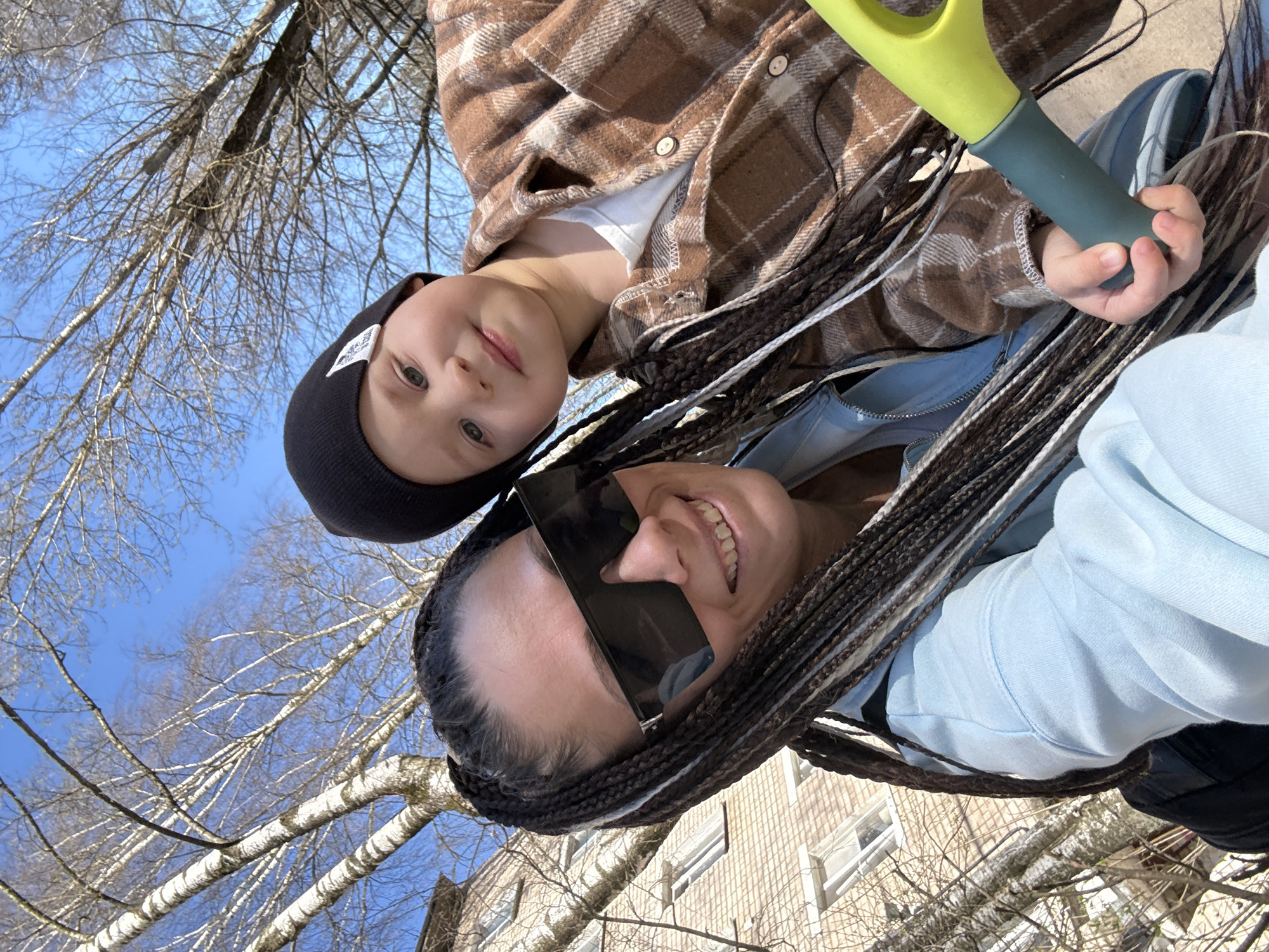 This outdoor portrait features two individuals, an adult woman and a young child, captured from a low angle looking upwards. The image is set against a bright, clear blue sky with numerous bare, leafless tree branches, indicating late winter or early spring in Rusanovo, Russia. A light-colored brick building with windows is visible in the lower background.
The adult woman, positioned lower in the frame, has long, dark braided hair with some lighter strands. She wears large black rectangular sunglasses and a light blue hooded sweatshirt, and is smiling broadly, revealing her teeth.
The child, positioned above and slightly to the left of the woman, has light-colored eyes and a neutral expression. They are wearing a black beanie with a small, unreadable white patch or logo, and a brown and white plaid shirt over a white t-shirt. The child is holding a green and grey toy shovel or spade in their right hand.
The scene conveys a casual and happy moment outdoors, with both subjects looking towards the camera, possibly for a selfie. The clear sky suggests a sunny day.