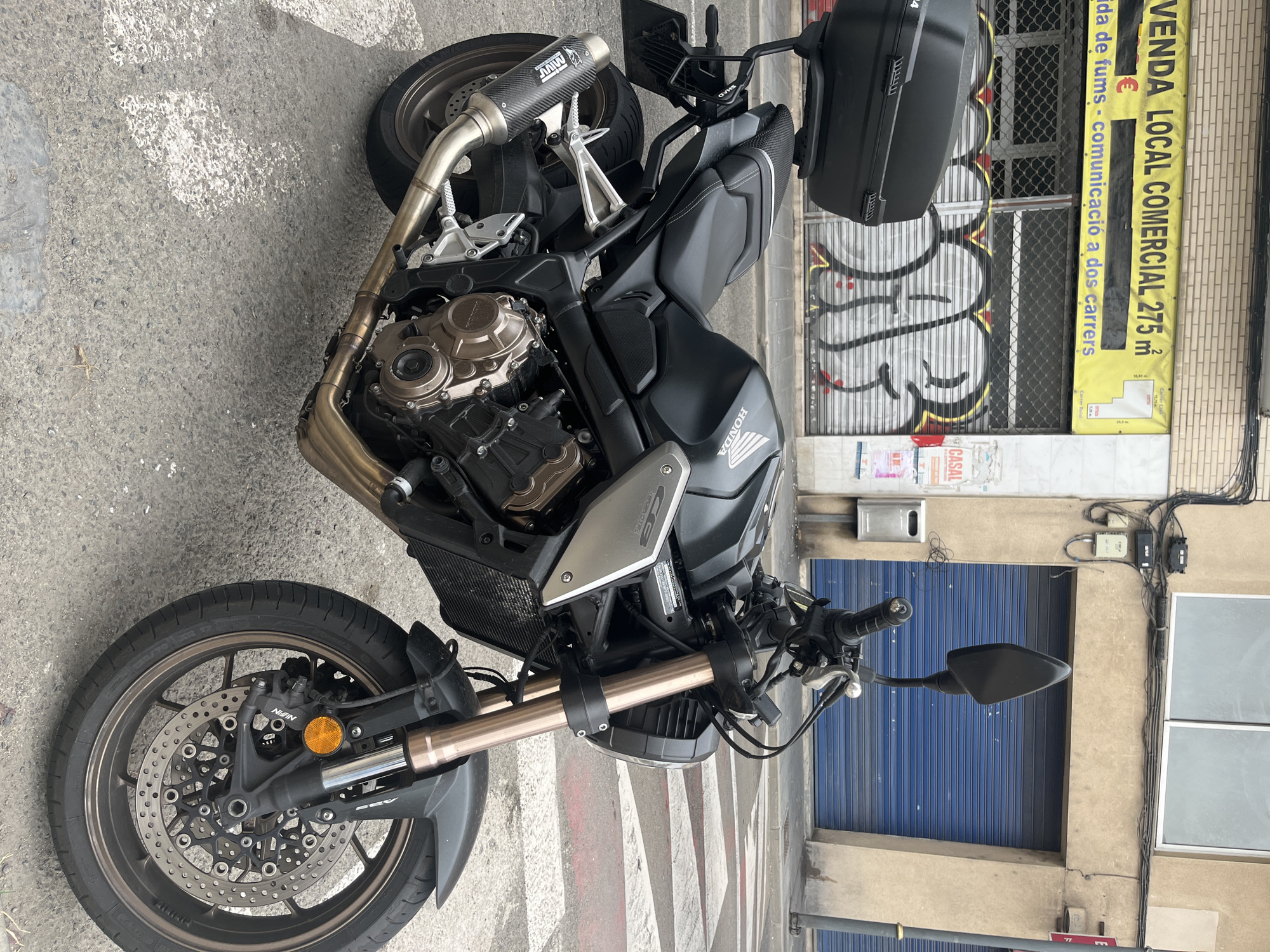 A black Honda motorcycle is parked on a pedestrian crossing on a street in Tarragona, Spain. The motorcycle has a black top box and a carbon fiber exhaust. In the background, there is a building with a yellow banner that reads "VENDA LOCAL COMERCIAL 275m²" and graffiti on the metal shutters. The building also has blue shutters and a street lamp. The sky is overcast.
