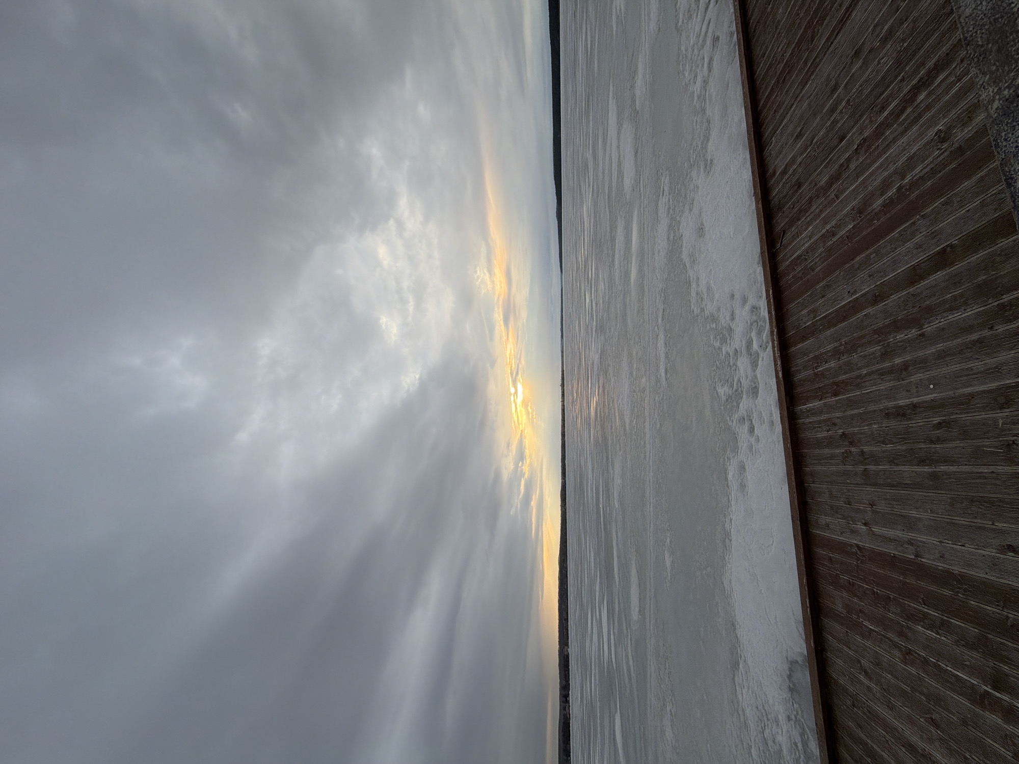 The image depicts a frozen lake adjacent to a wooden boardwalk or deck in Votkinsk, Russia. The scene is oriented horizontally, with the sky on the left side. The sky is overcast, with a small break near the horizon allowing sunlight to pierce through. The frozen lake has a mottled, textured appearance with visible cracks and patterns on its surface. The boardwalk is made of parallel planks of wood and appears to be adjacent to the lake’s frozen edge. There are no people visible.