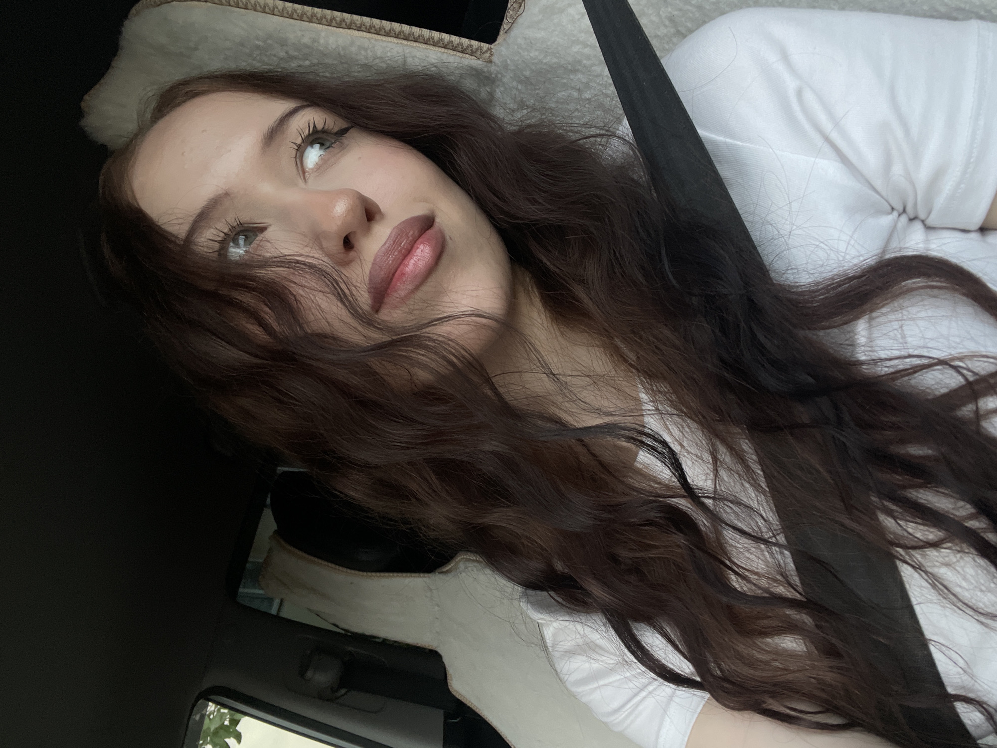 A young woman with long, wavy brown hair is in the passenger seat of a car, looking to her right. She is wearing a white t-shirt and a black seatbelt is across her chest. The headrest behind her is covered in a beige, fuzzy material. The interior of the car is visible, including the dashboard and a portion of the exterior through the window. The scene appears to be taking place in Votkinsk, Russia, though no specific landmarks identifying the city are visible within the frame.