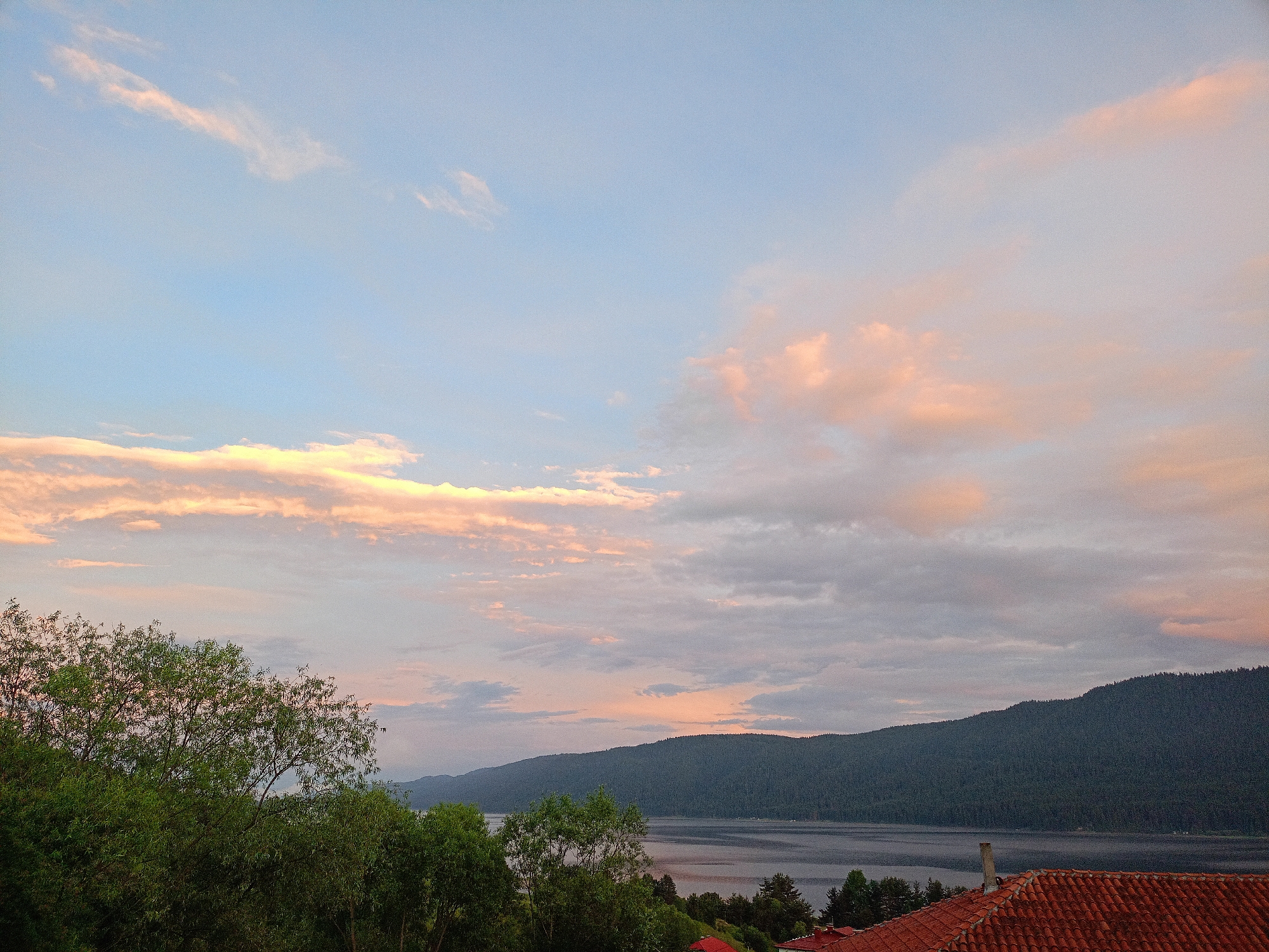 The image depicts a serene natural landscape at dusk, likely taken from an elevated position in Sarnitsa, Bulgaria. The foreground features lush green trees and the terracotta-tiled roof of a building. Beyond this, a body of water, possibly a lake, stretches out, bordered by a heavily forested mountain range. The sky is a gradient of soft blues and pinks, with scattered clouds catching the fading sunlight, suggesting it is either sunrise or sunset. The overall impression is one of tranquility and natural beauty. There are no people or animals visible in the frame.
