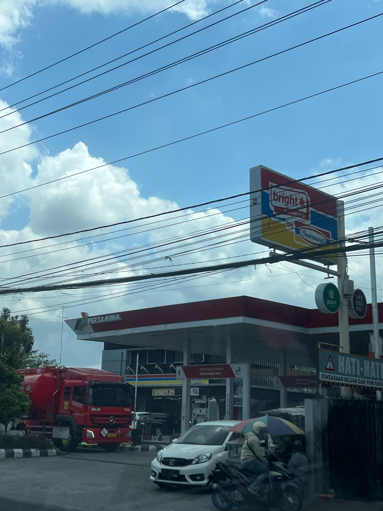 This image depicts a street scene in Magelang, Indonesia, centered around a Pertamina gas station. The sky is bright blue with scattered white clouds, and several power lines are strung across the upper portion of the frame.
Dominating the mid-ground is the red and white Pertamina gas station canopy. To the right of the canopy, a large, illuminated sign reads "bright" with smaller text below it that says "by Indomaret." Below this sign, on a pole, are circular green and black signs, one with the text "POINT" and a green circle with a white dot.
In the foreground, a red tanker truck is parked to the left. The truck has a Mercedes-Benz emblem on its grille. To the right of the truck, a white car is visible. Further to the right, two people are on a motorcycle, one wearing a helmet and holding an umbrella. A black metal fence is positioned behind them.
The ground is paved, with a striped pedestrian crossing in front of the tanker truck. The overall impression is a busy urban environment with commercial activity.