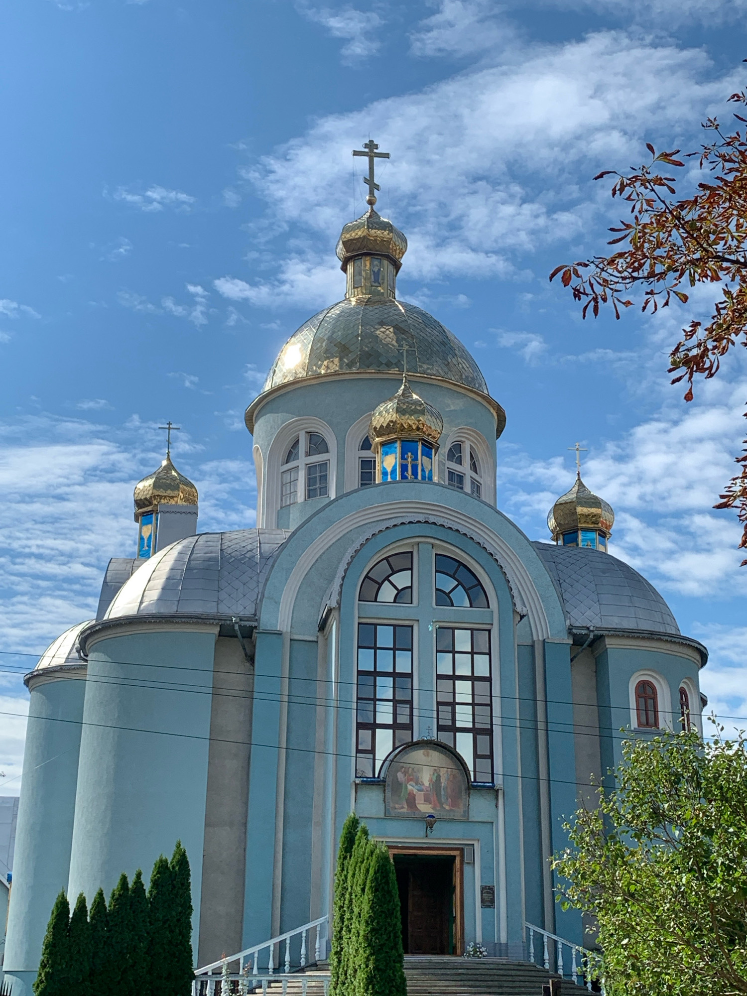 An exterior, low-angle, full shot captures the facade of a blue and grey Eastern Orthodox church in Kolomyia, Ukraine, on a partly cloudy day. The church features multiple golden domes topped with crosses, a large arched window with multiple panes above the entrance, and a fresco depicting a religious scene above the doorway. A set of stairs leads up to the open entrance. Cypress trees flank the stairs, and a deciduous tree with reddish-brown leaves is visible in the upper right corner. Power lines are strung across the front of the building.