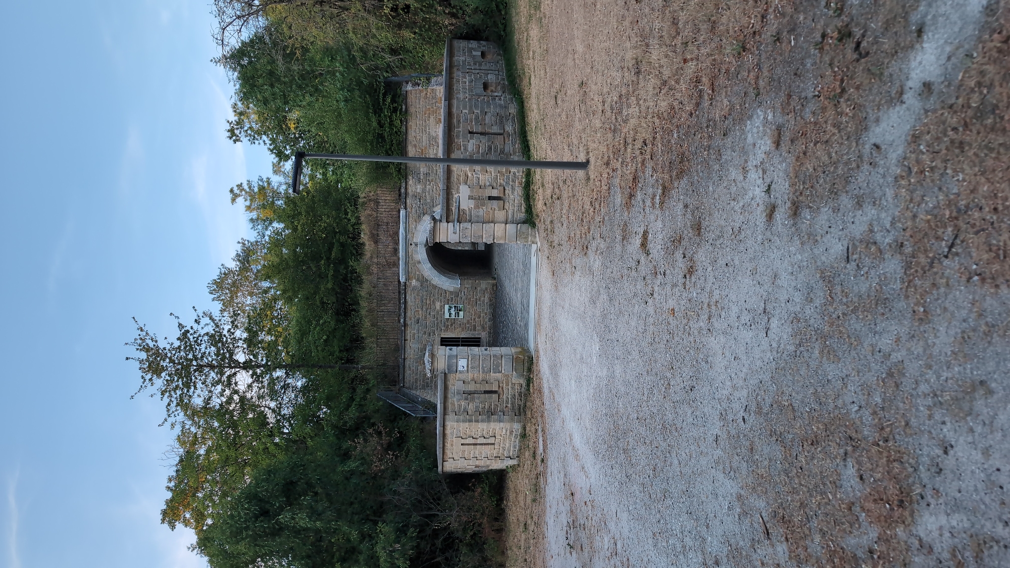 The image shows a view of an old stone fort entrance with a gravel path leading up to it. The fort has a large arched opening and is flanked by stone walls with decorative elements. There is a modern street lamp to the right of the entrance. The surrounding area is covered with dry grass and trees, suggesting an outdoor, possibly rural or park-like setting. The sky is clear blue with a few wispy clouds. There are no people visible in the image. The architecture of the fort suggests a historical military structure.
