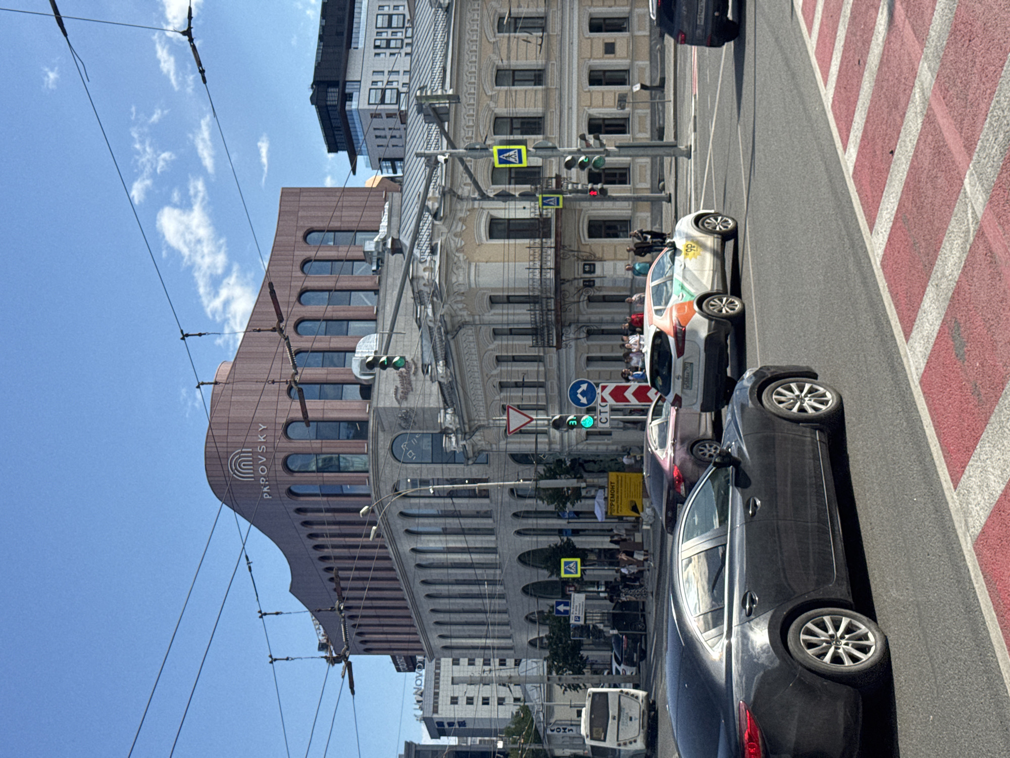 This is a daytime, eye-level shot of a street scene in Yekaterinburg, Russia, captured from an urban intersection. The foreground is dominated by a black car in the lower left, and red and white pedestrian crossing markings. Beyond this, a white bus is visible on the left, and further to the right, several cars are in motion or stopped at the traffic lights, including a white and orange car with a logo on its side. The middle ground features a row of buildings, with a striking curved, terracotta-colored building with arched windows and the text "PAROVSKY" visible on its upper facade. To the right of this is a more ornate, older building with classical architectural elements. The background shows more modern high-rise buildings. Traffic lights, street signs, and overhead power lines are prevalent throughout the scene. A group of pedestrians are gathered on the sidewalk, some near a bus stop and others near one of the buildings. The sky is bright blue with scattered white clouds.