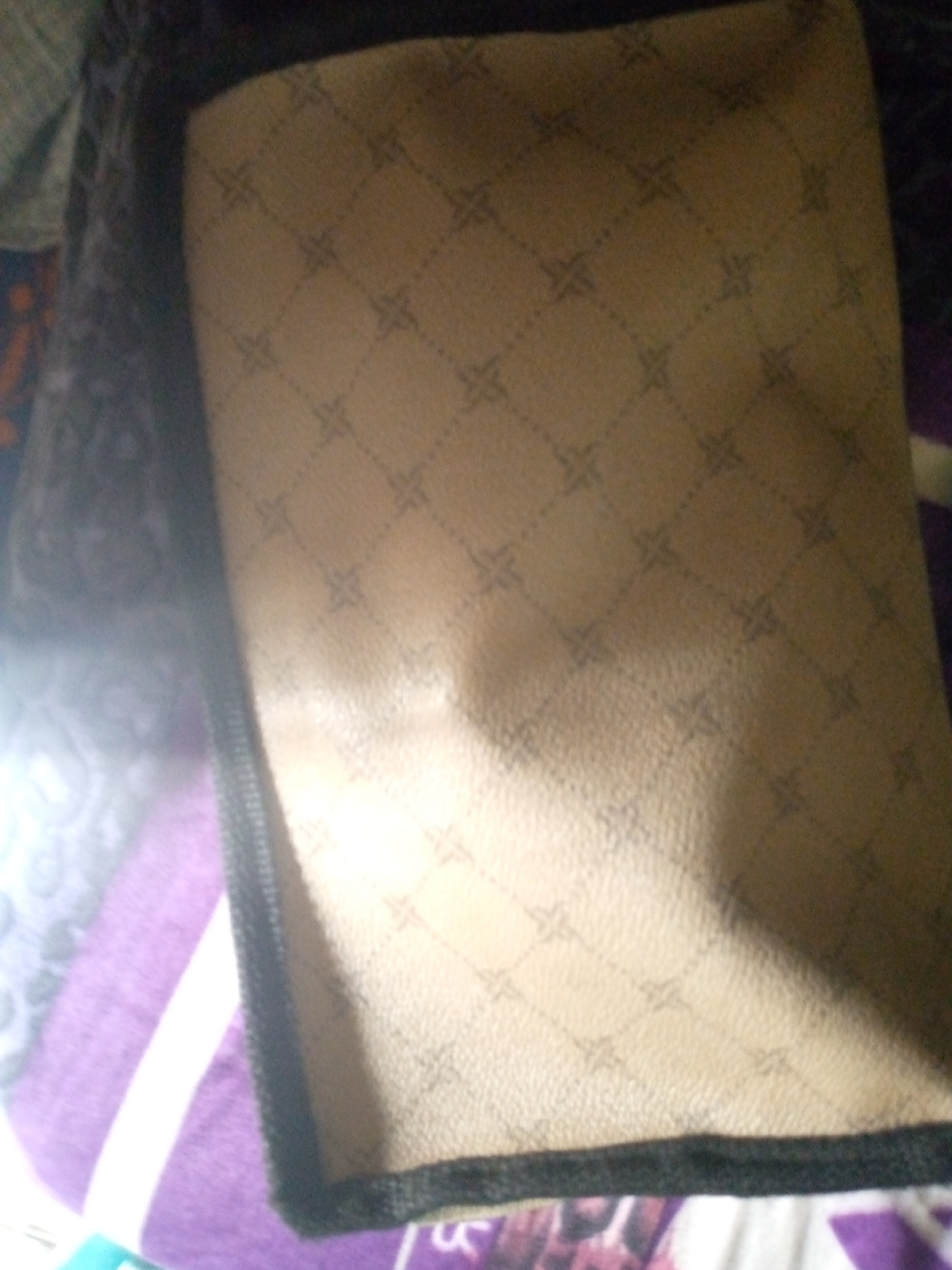 The image shows a close-up, angled view of a tan-colored fabric with a repeating diamond pattern. The fabric appears to be some sort of accessory or bag, with a dark border along the bottom edge. The lighting suggests it is indoors, and the background is blurry, showing hints of purple and white fabric. The exact location within Jalingo, Nigeria, is not discernible from the visual information provided.