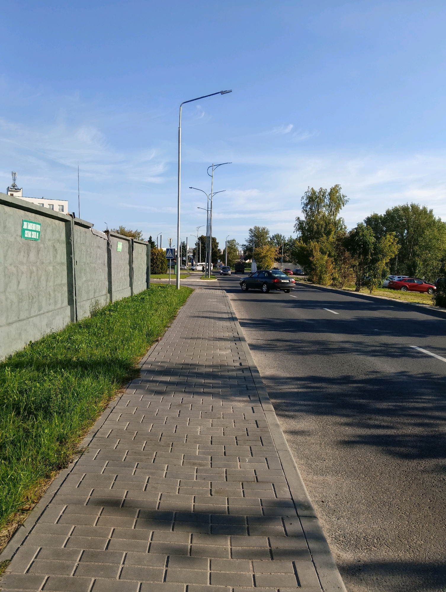 The image depicts a sunny daytime scene on a street in Hrodna, Belarus. On the left, a grey concrete wall runs along the edge of a grassy area. The wall has a green rectangular sign attached to it. A paved sidewalk, composed of grey rectangular blocks, runs parallel to the wall. To the right of the sidewalk is a two-lane asphalt road with a white line marking the lanes. Several cars are visible on the road, with a dark sedan in the foreground and a red car parked on the right side. The street is lined with trees and lampposts. The sky is blue with some wispy white clouds. In the background, buildings and more trees are visible.